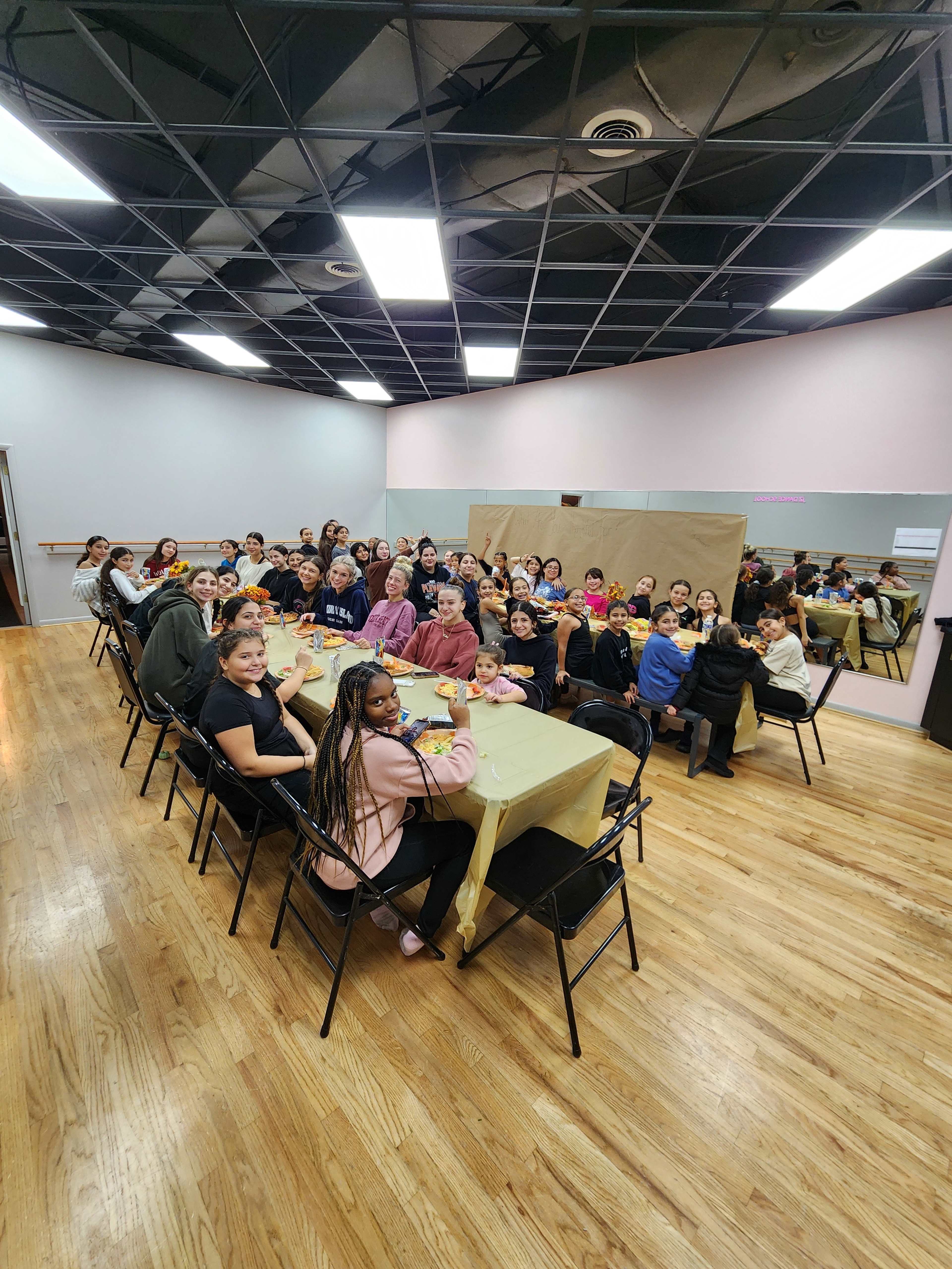 A large group of people sits around a long table in a spacious room with mirrors, all facing the camera.