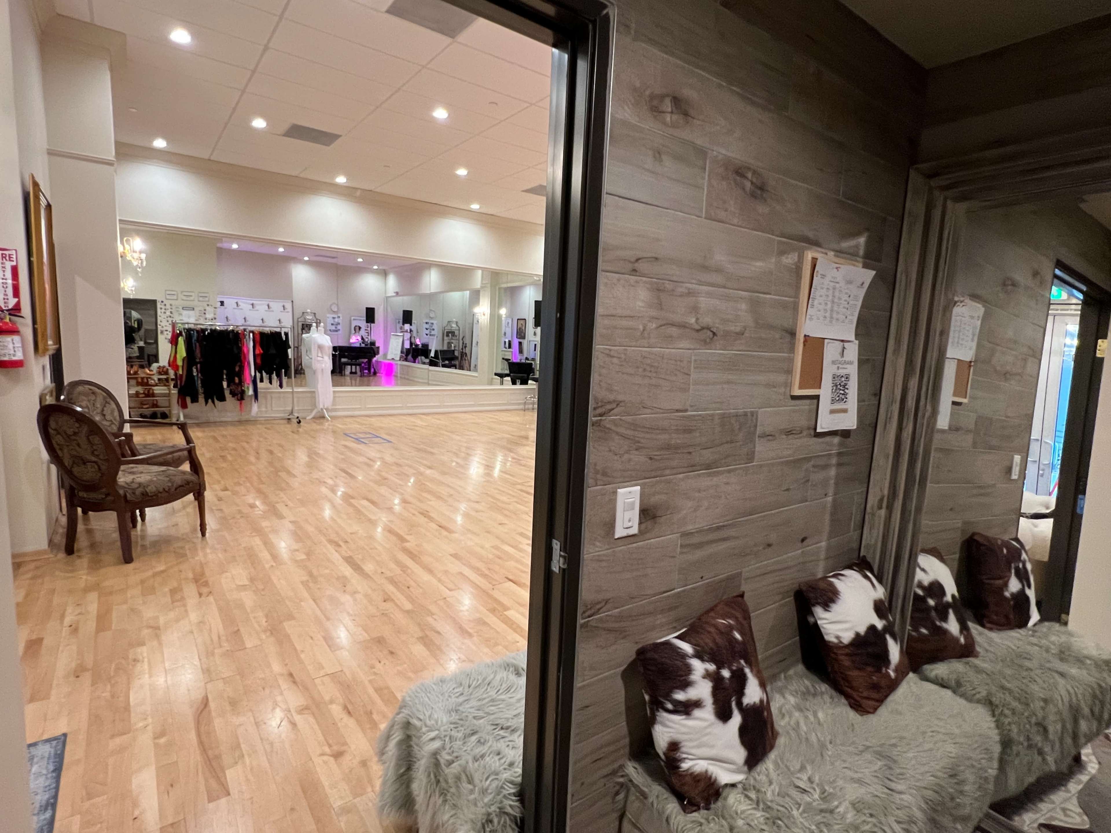 A view from a hallway into a dance studio with wooden flooring and a mirrored wall, featuring various dance attire hanging on a rack.