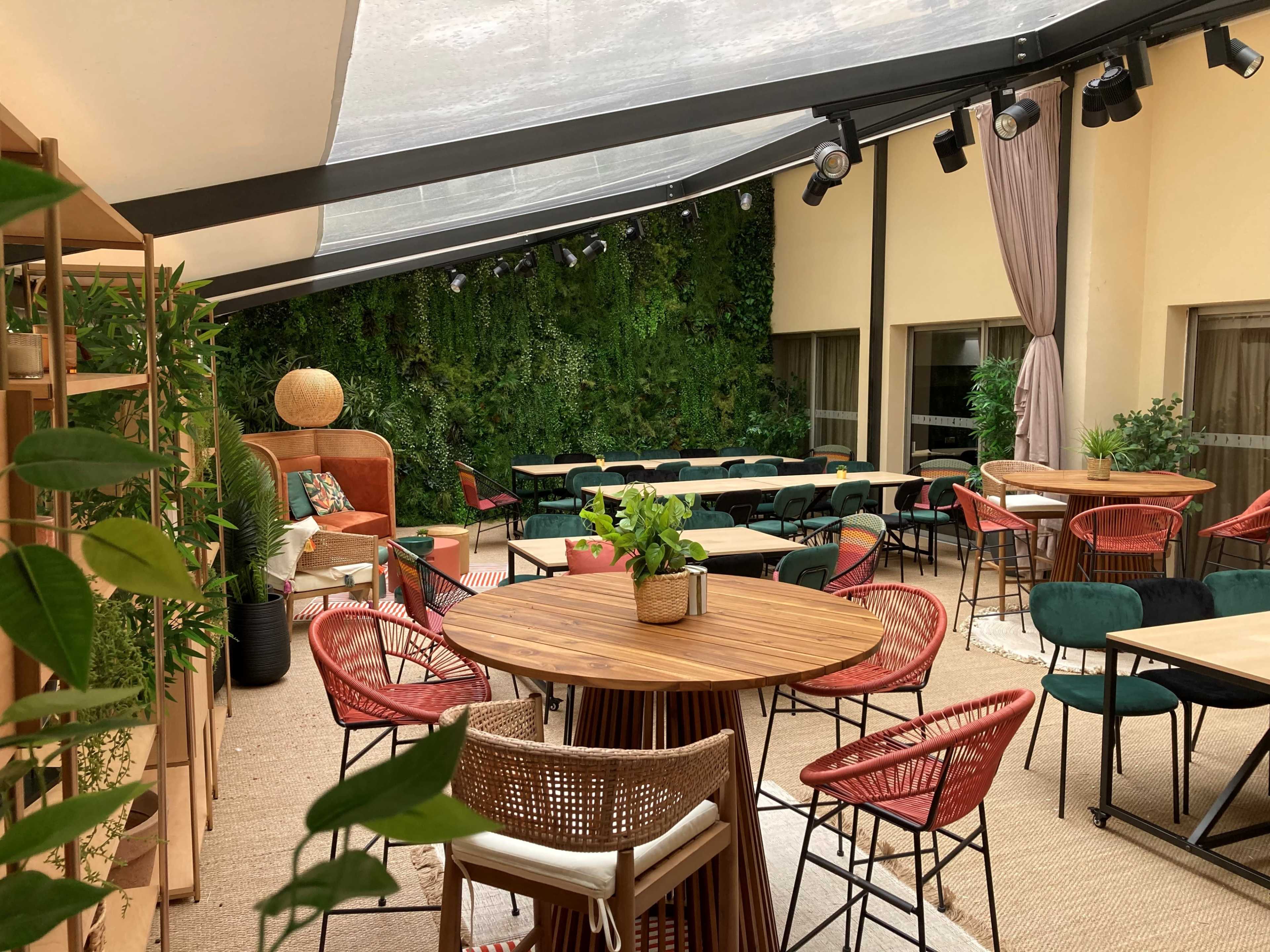 The image shows a stylish indoor-outdoor lounge area featuring wooden tables surrounded by colorful chairs, with a green wall of plants in the background under a transparent roof.