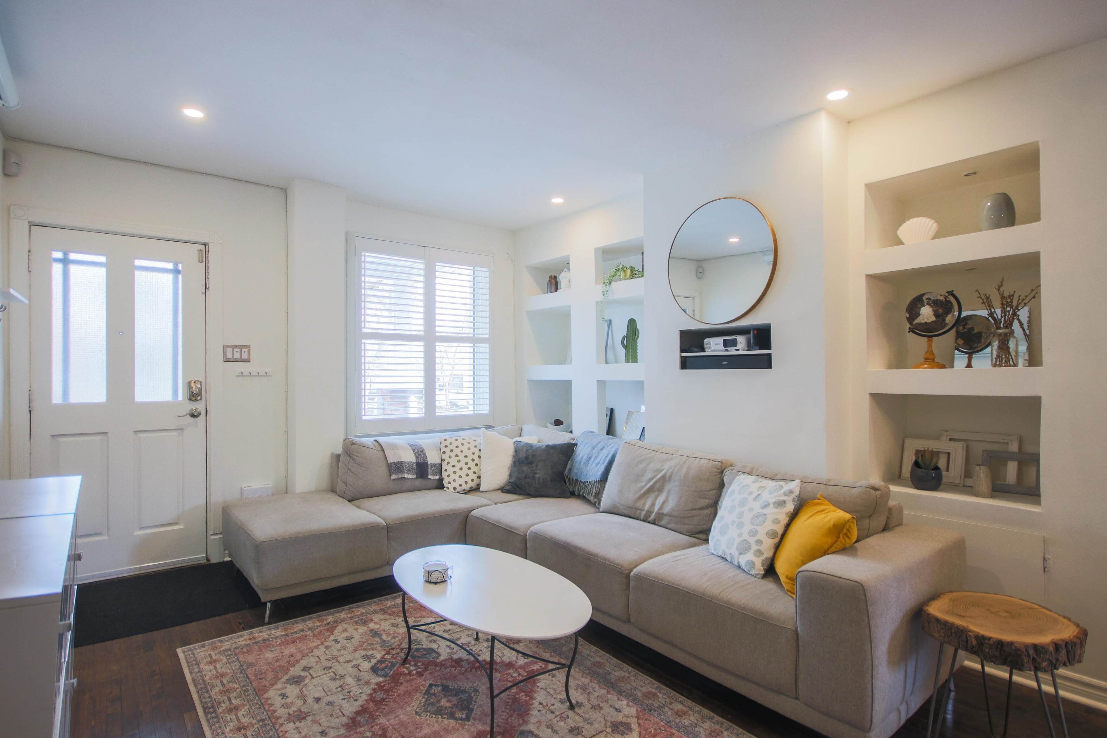 A modern living room features a sectional sofa, a round mirror, and built-in shelves, with a small coffee table and a decorative rug.