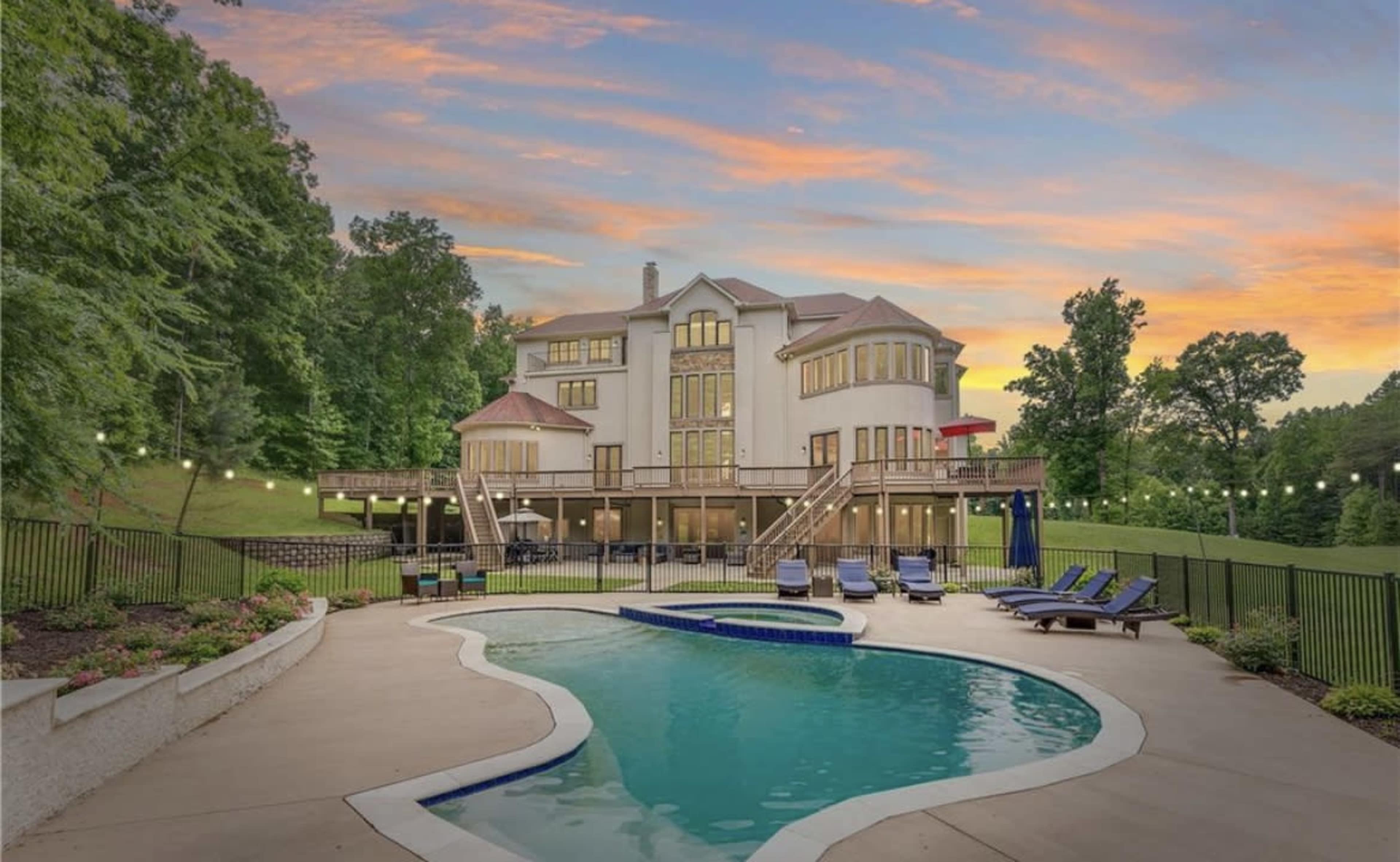 A large house with multiple balconies overlooks a swimming pool surrounded by lounge chairs at sunset.