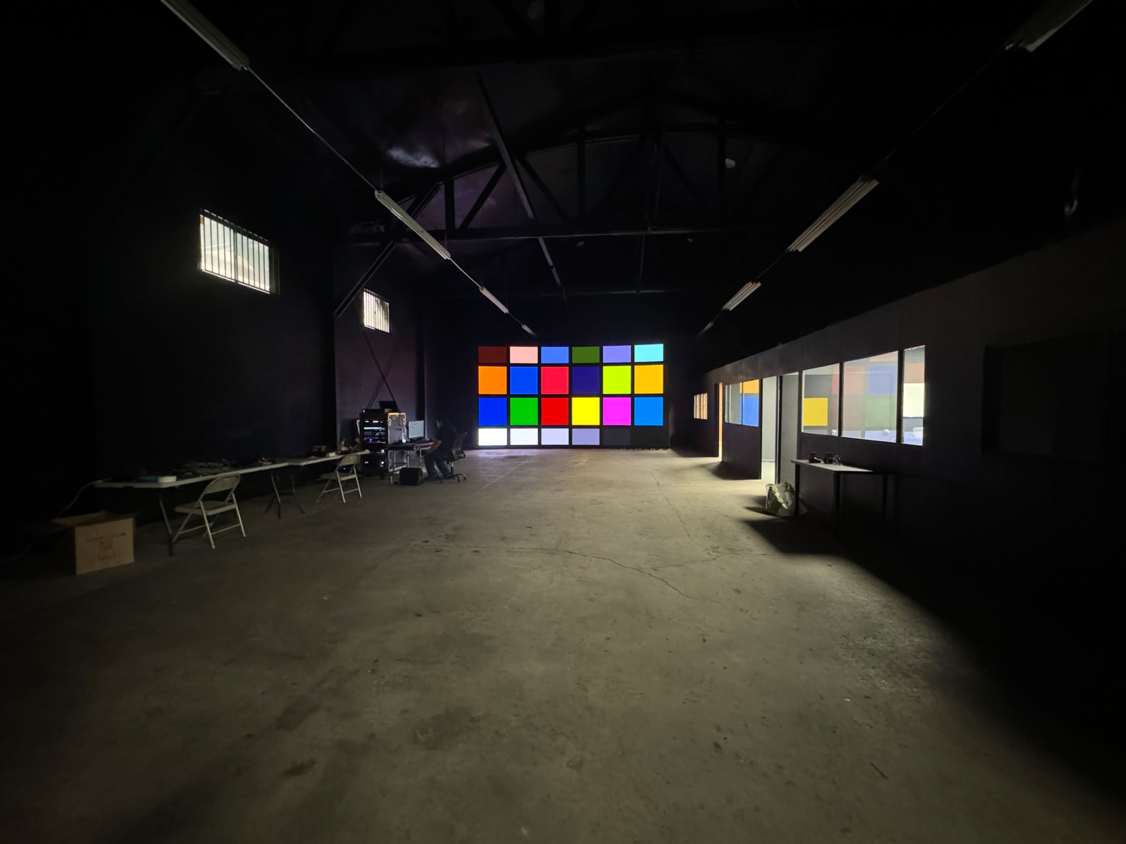 The image shows a large, empty warehouse space with a colorful grid display on one wall and a table with chairs on the left side.