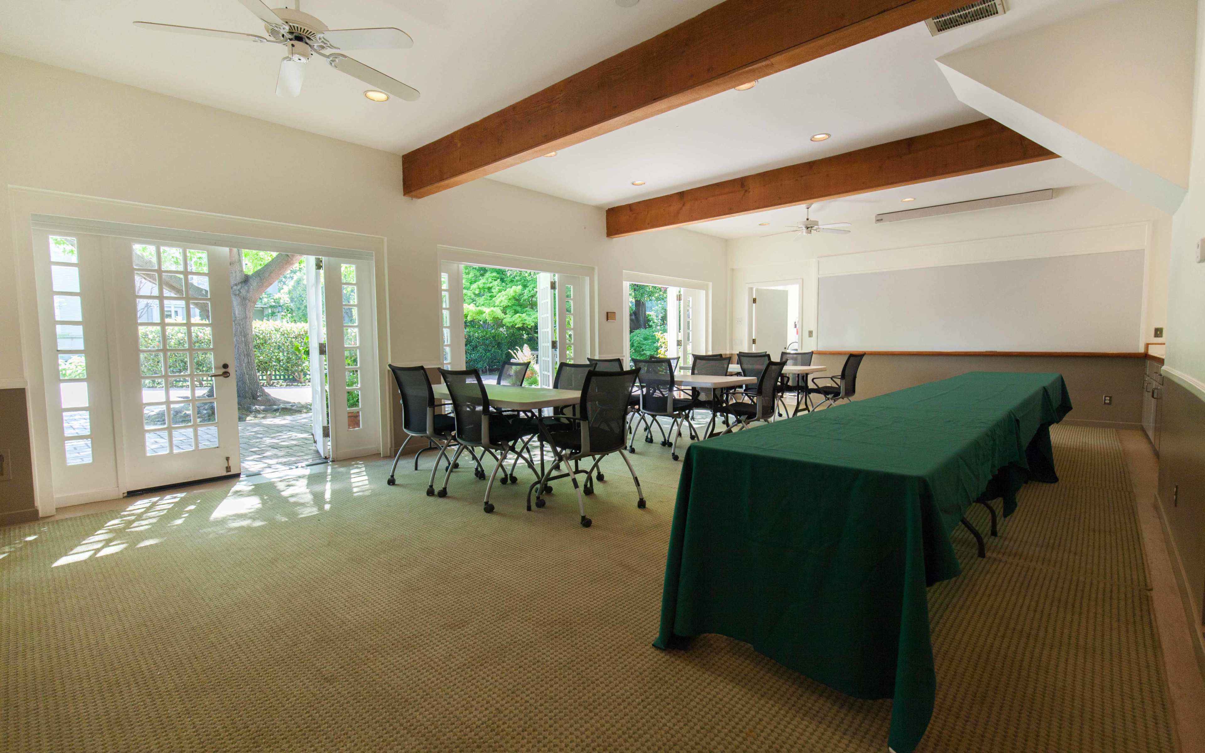 A conference room features a long table covered with a green tablecloth and several black chairs arranged around it, with large windows letting in natural light.