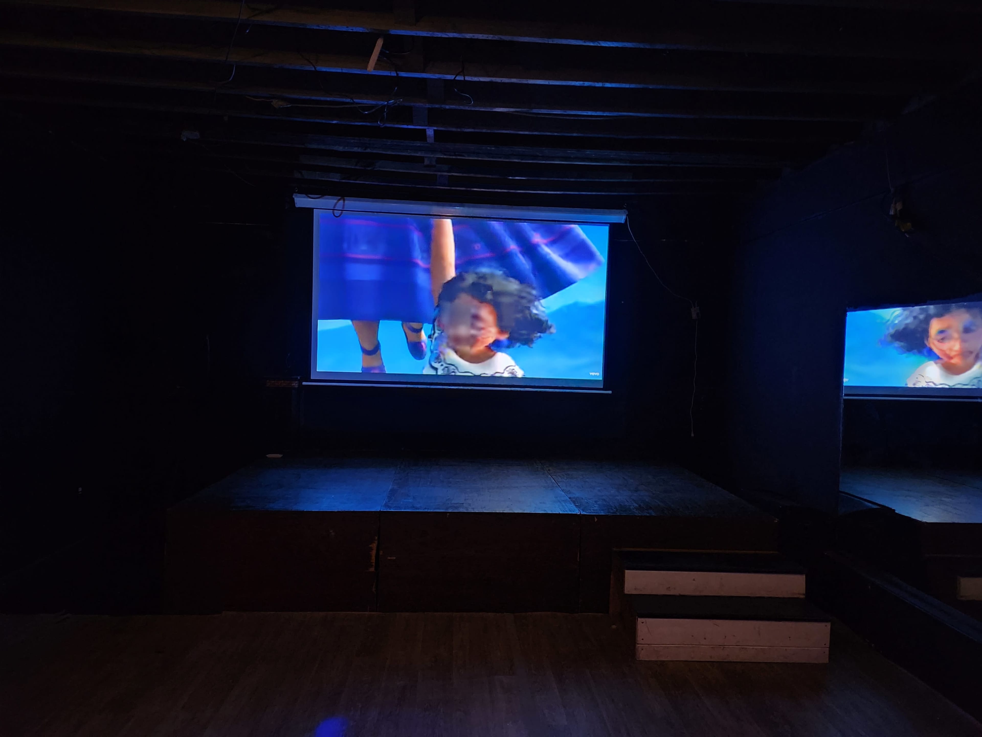 The image shows a dark room with a large screen displaying an animated scene, while a smaller screen mirrors the same image on an adjacent wall.