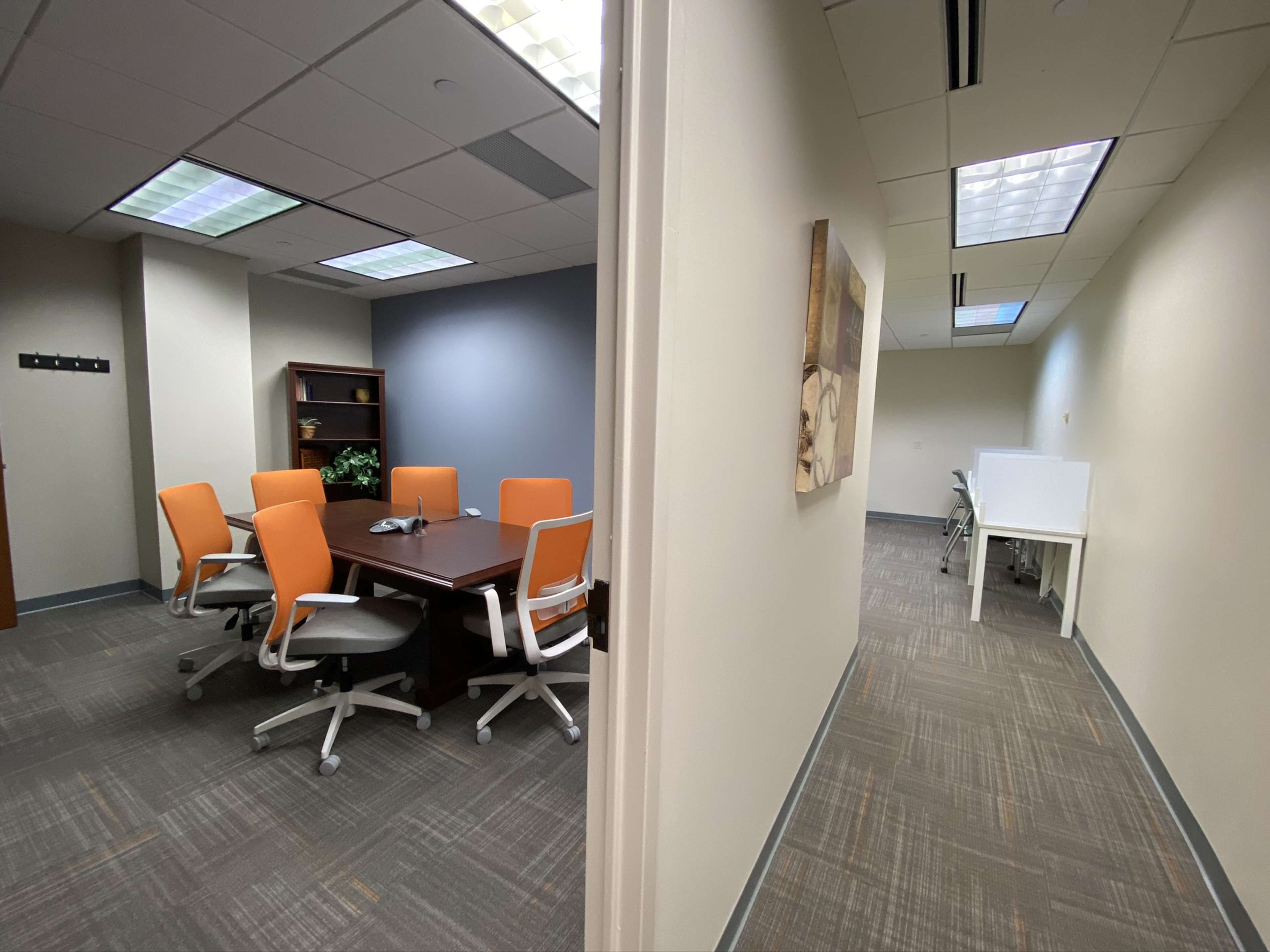 The image shows a divided office space featuring a meeting room with a wooden table surrounded by orange chairs on one side, and a hallway leading to another workspace on the other side.