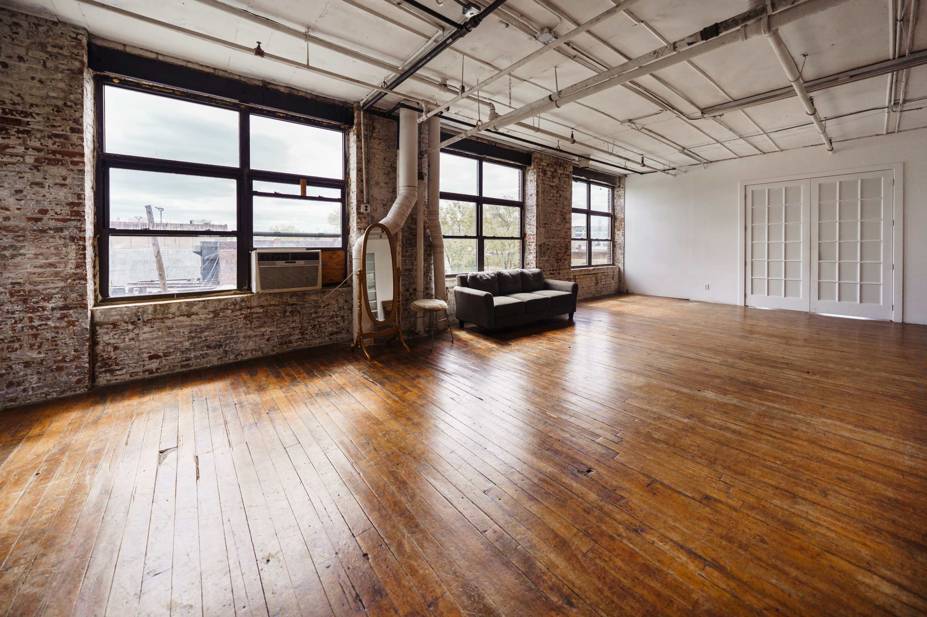 A spacious room with large windows, exposed brick walls, and hardwood flooring, furnished with a sofa and a decorative mirror.