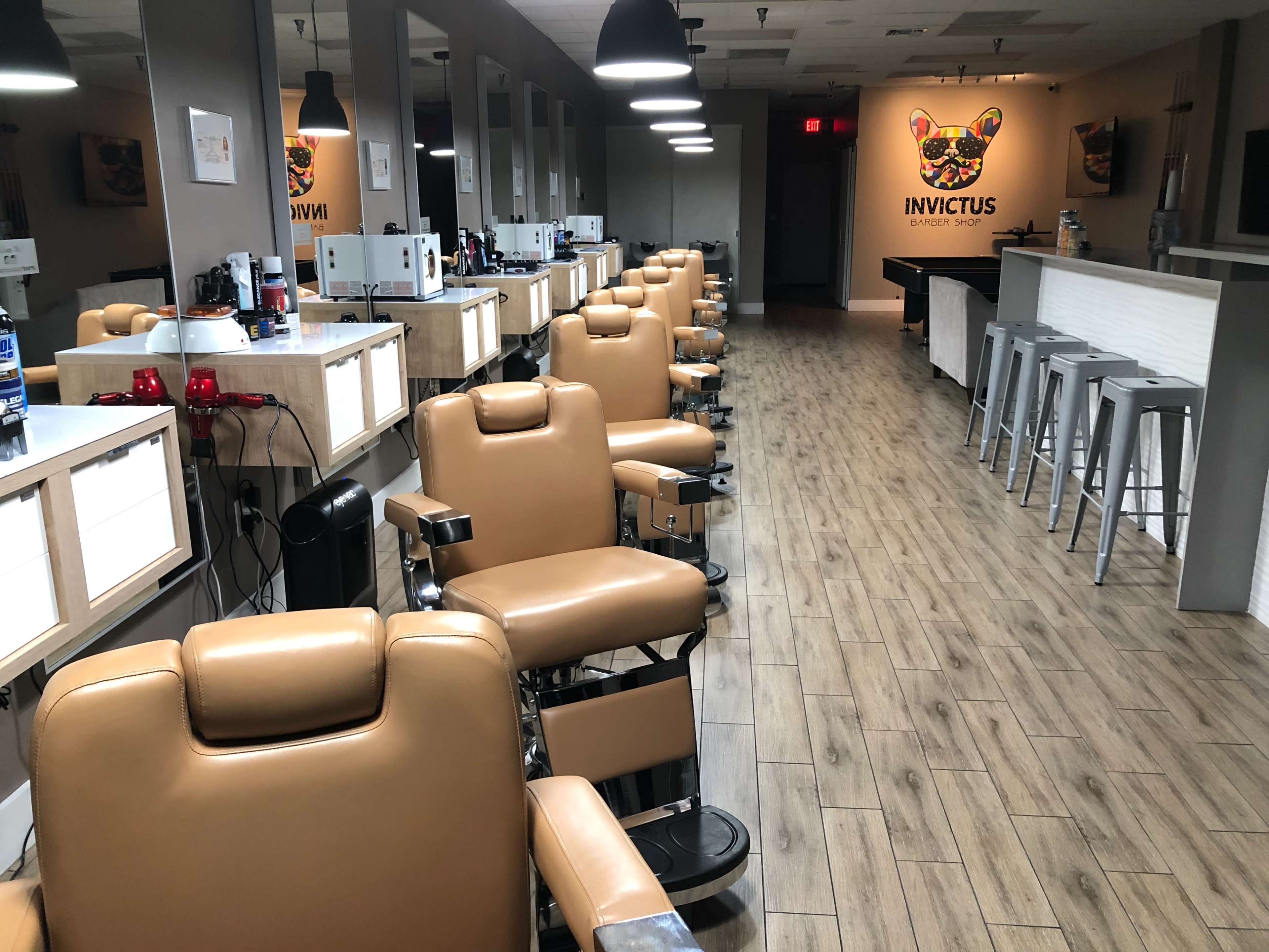 A modern barbershop with several beige barber chairs lined up in front of brightly lit mirrors and a prominent "INVICTUS" sign on the wall.
