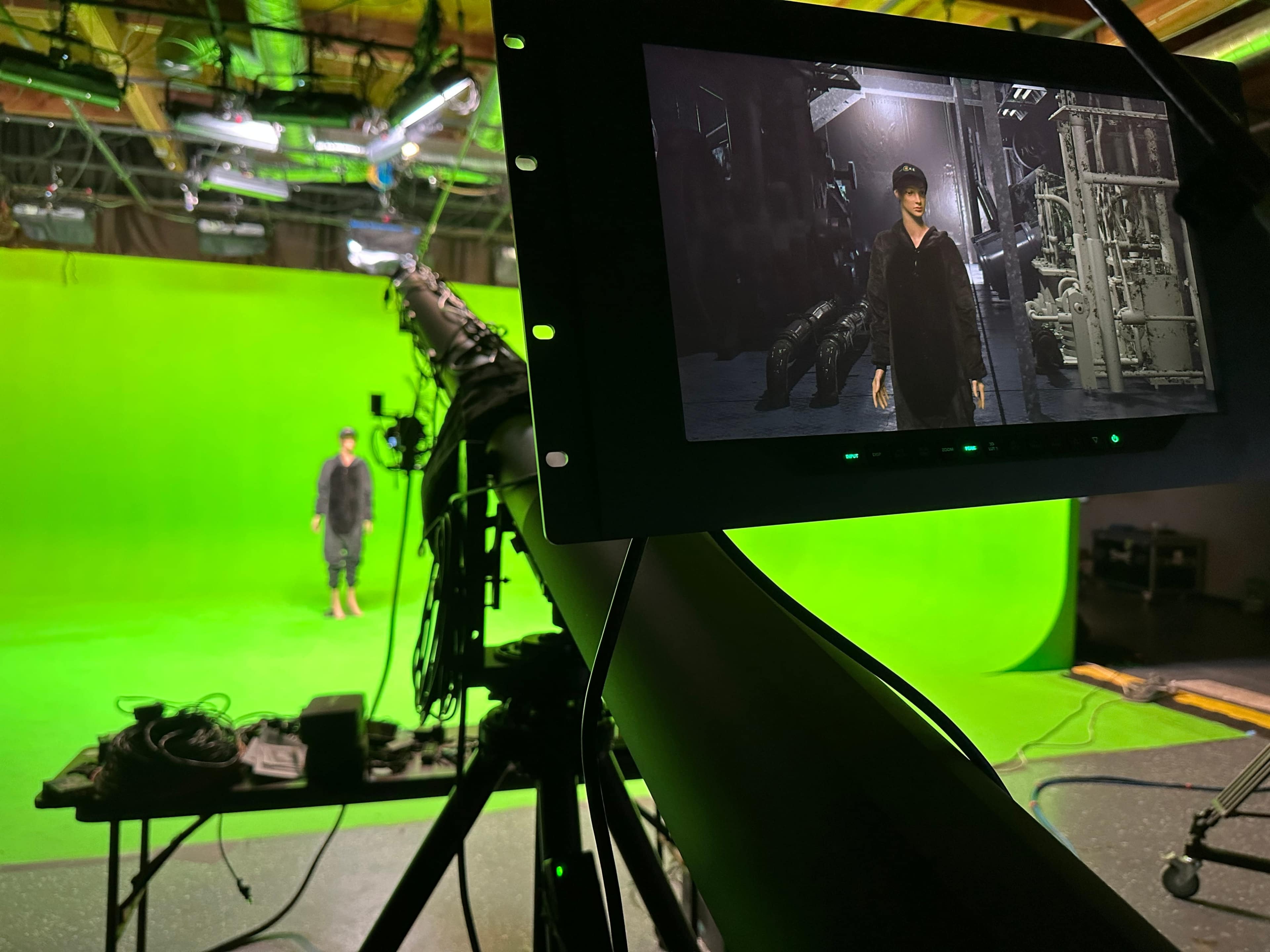 A person stands in front of a green screen inside a studio, while a camera and equipment are set up for recording or broadcasting.