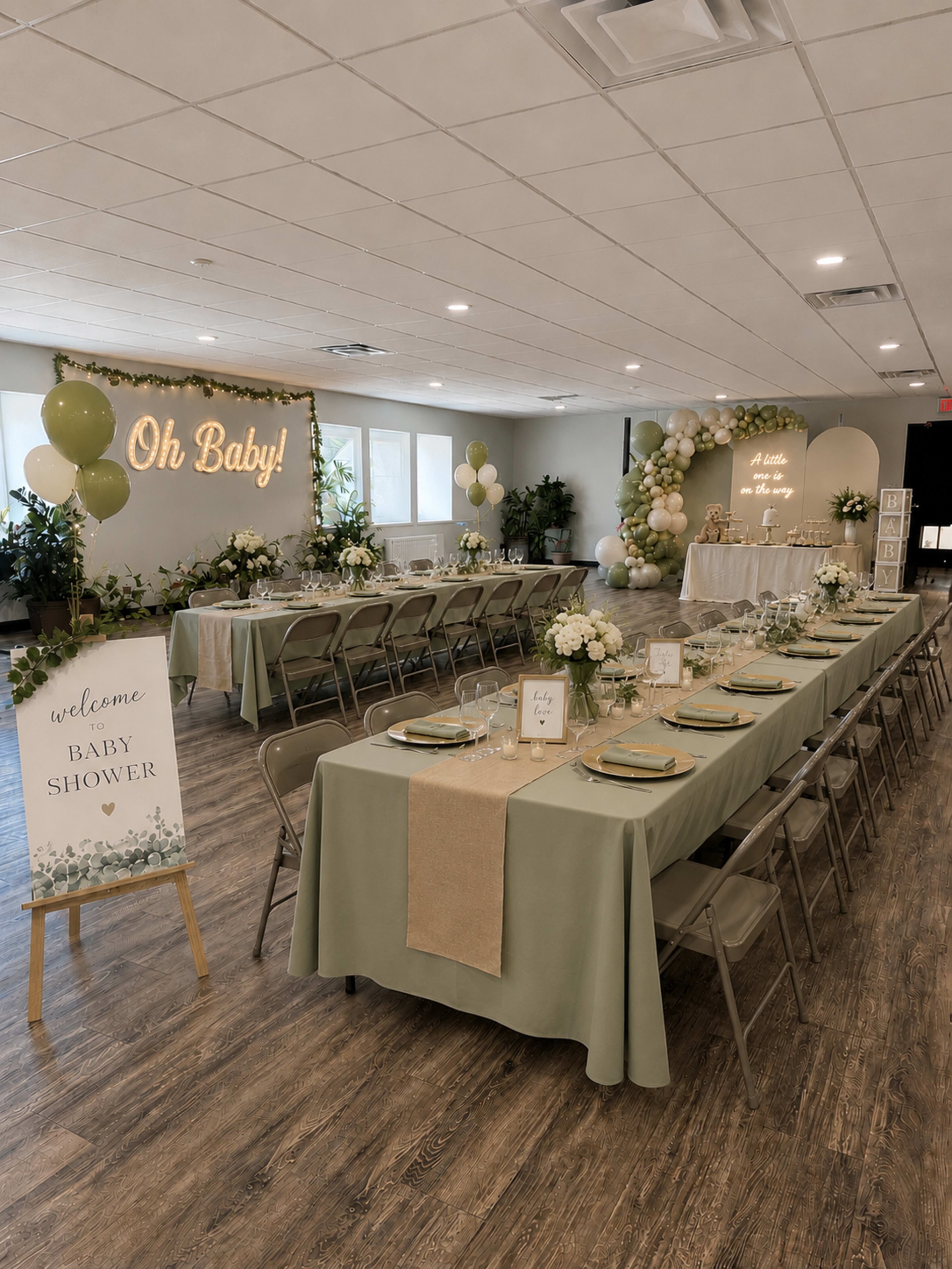 A spacious indoor venue is decorated for a baby shower, featuring tables with green tablecloths, flower centerpieces, and a large backdrop with the sign "Oh Baby!" along with balloon arrangements and a welcome sign.