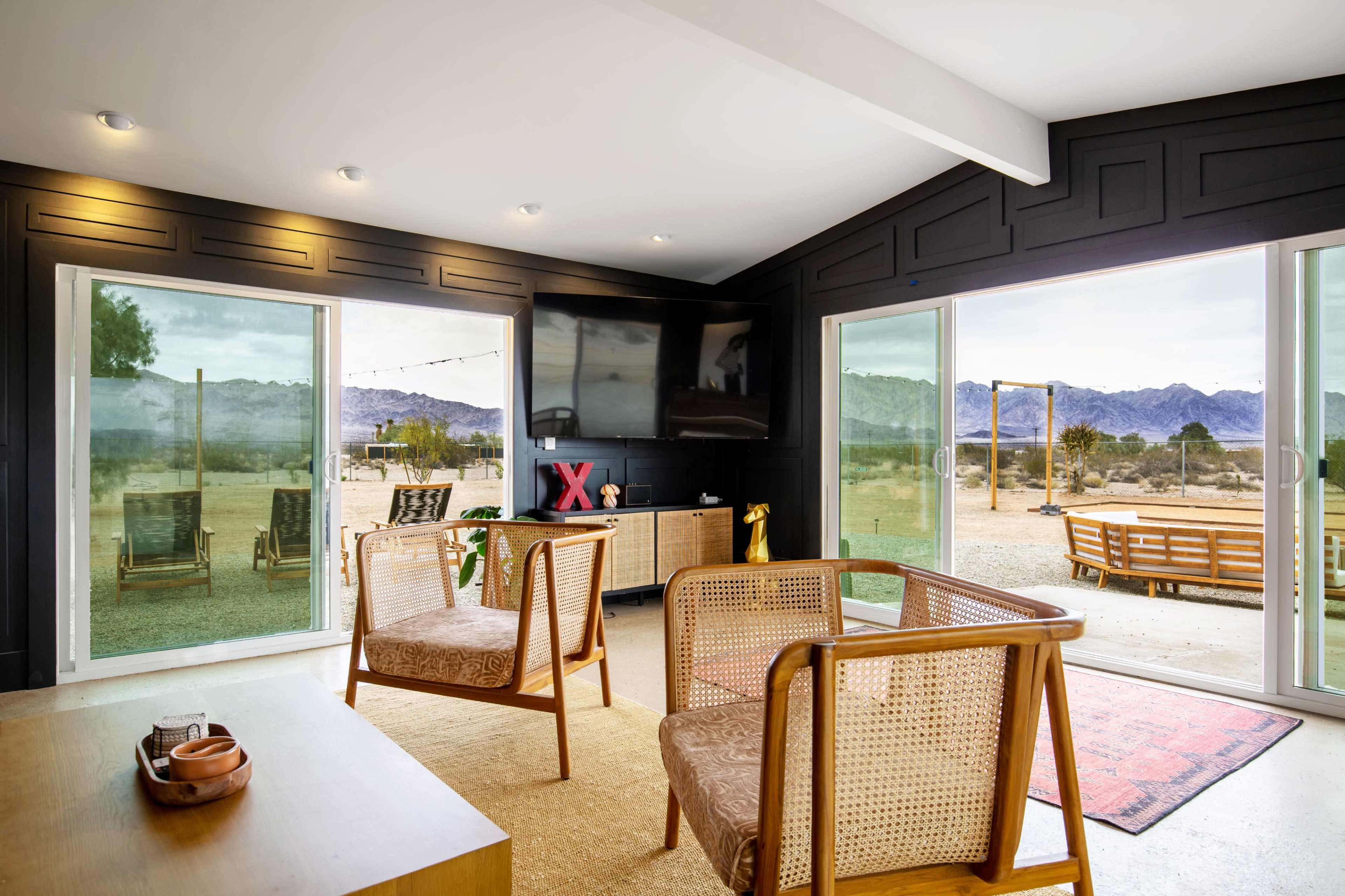 The image shows a modern interior living space with large windows overlooking a desert landscape, featuring wicker chairs and a television.