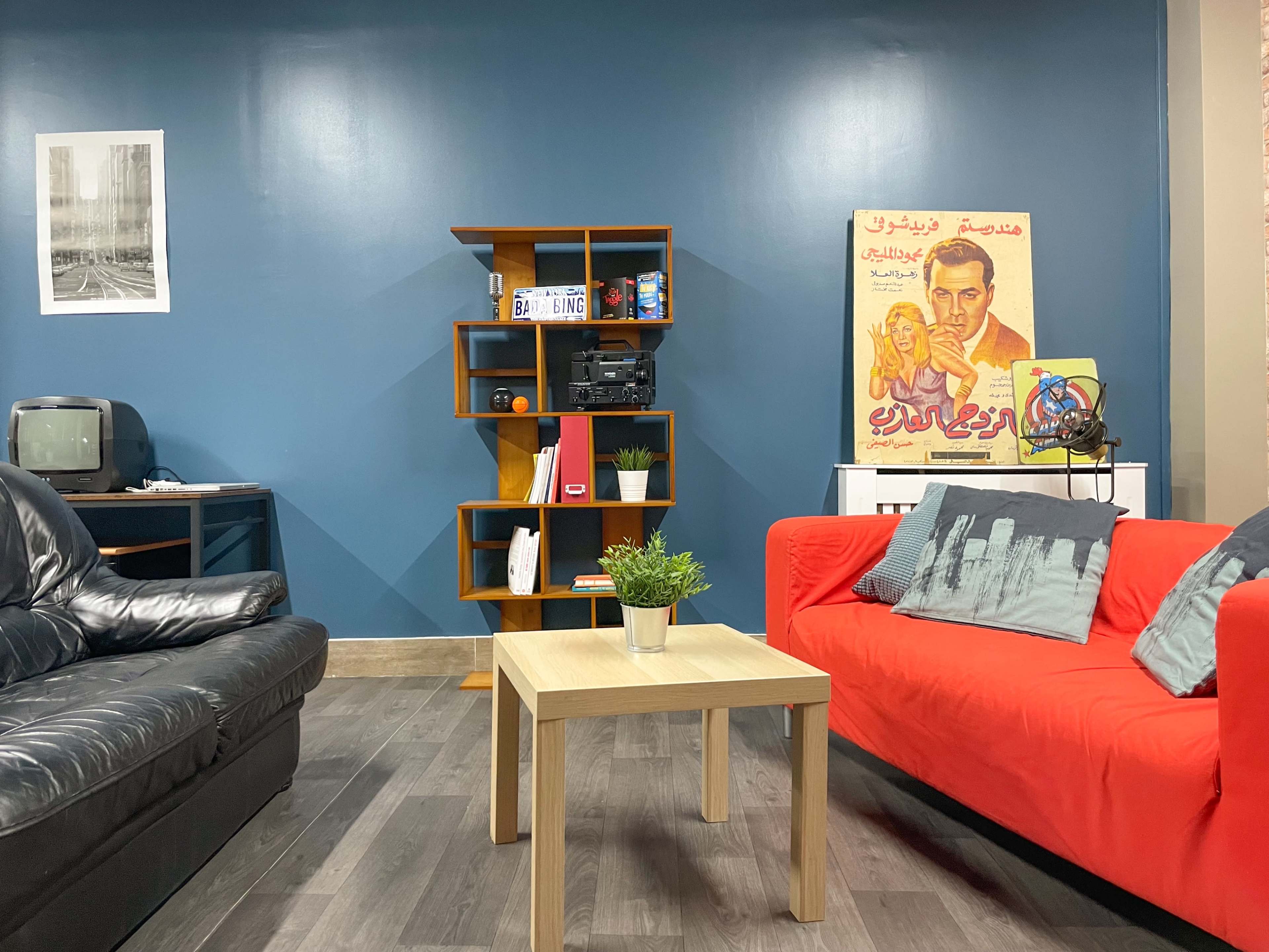 A living room with a black leather couch, a red fabric couch, a wooden coffee table, a bookshelf with various items, and a wall adorned with a vintage poster and a framed photograph.