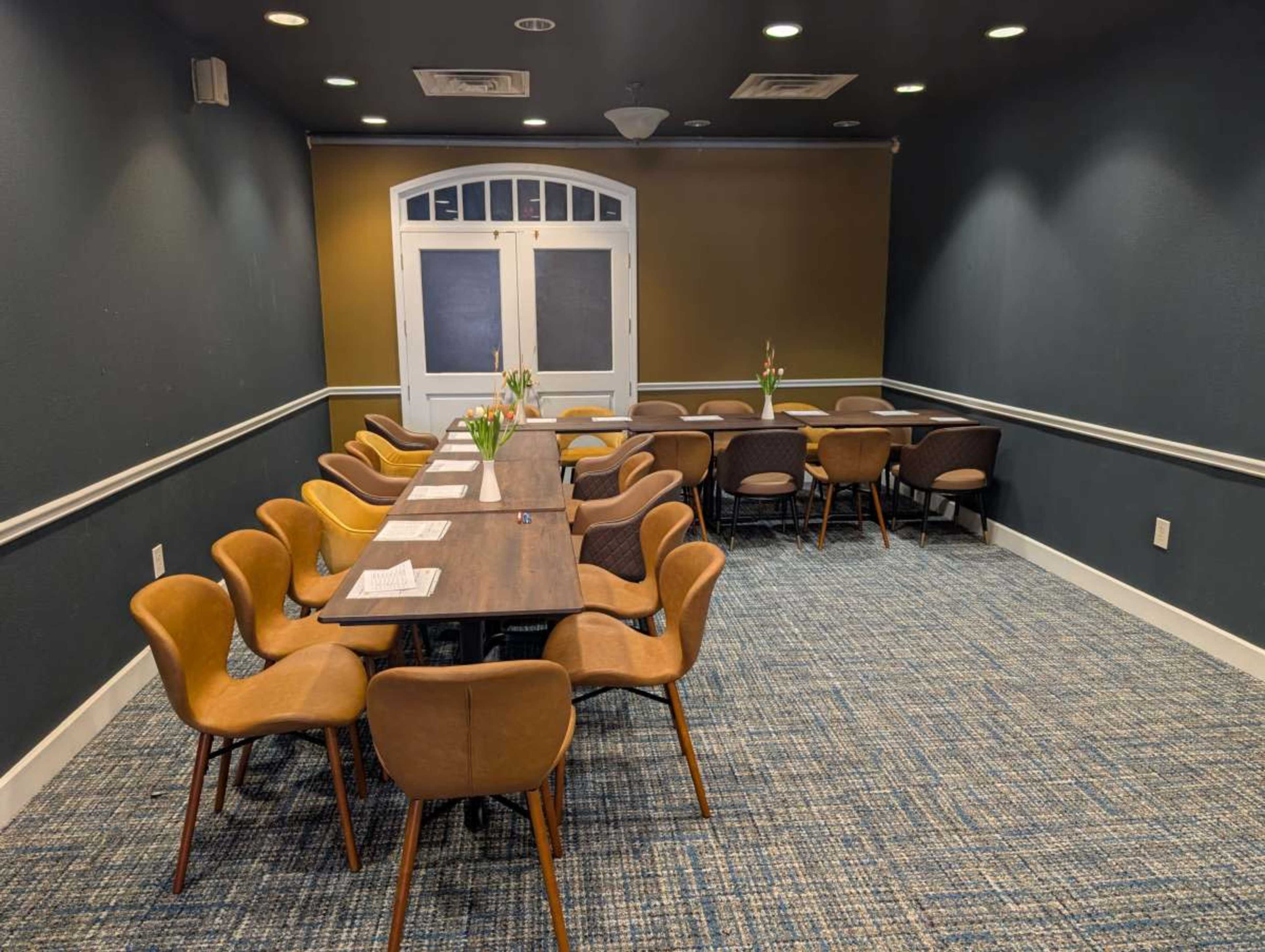 A long wooden table with seating surrounds a small meeting room featuring dark walls and a carpeted floor.