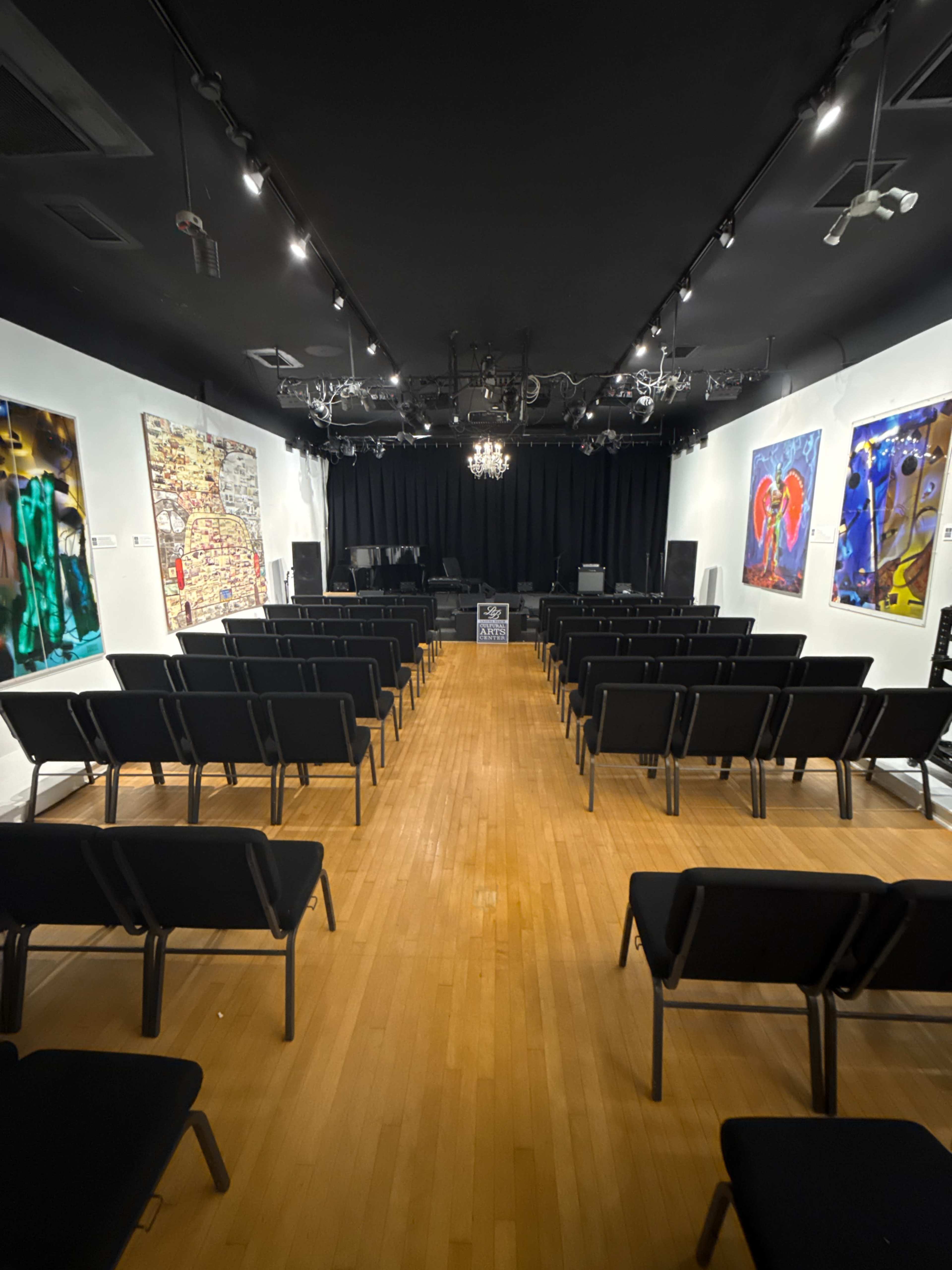 The image shows a performance space with rows of black chairs facing a stage, adorned with colorful artworks on the walls.