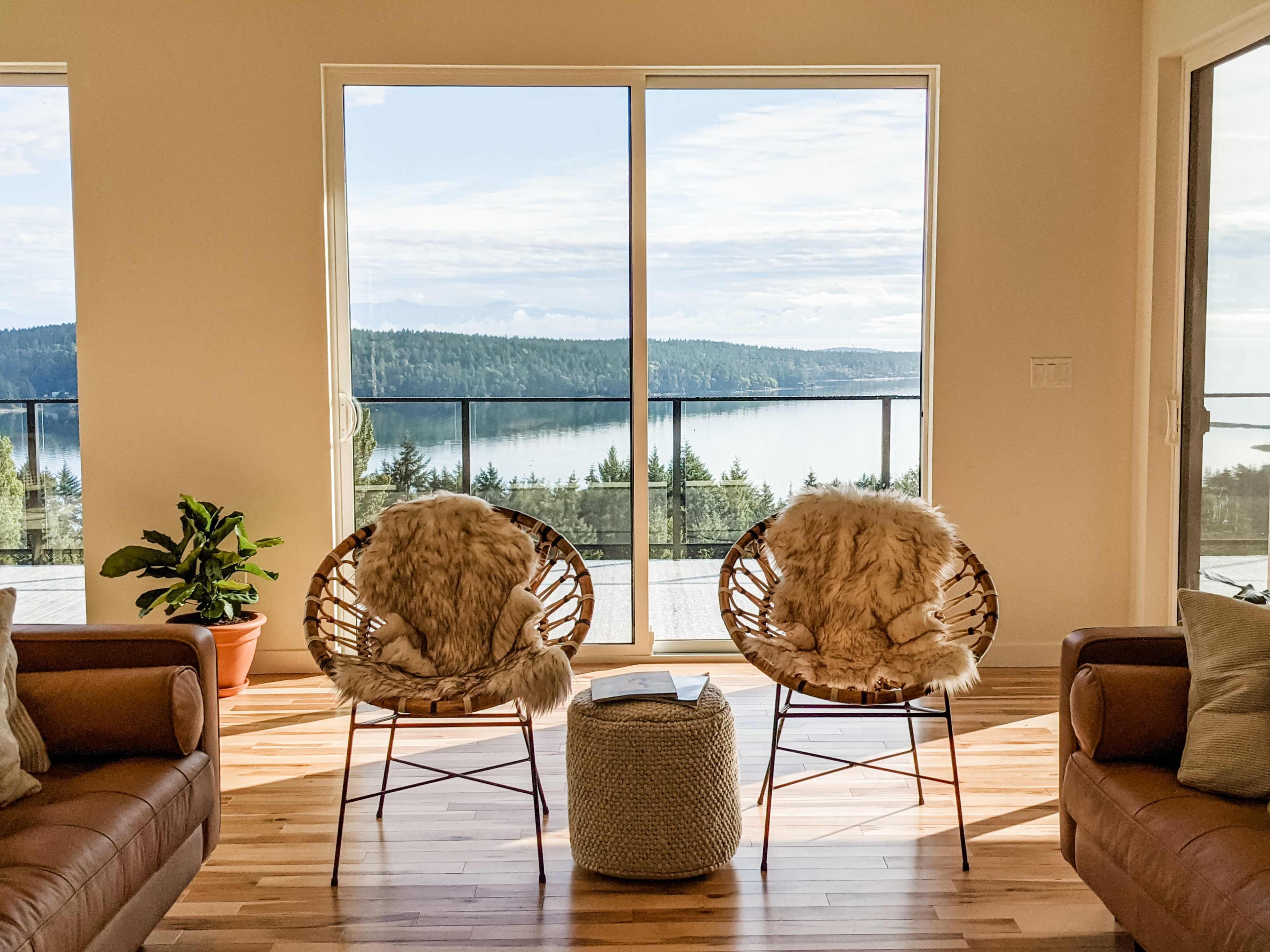 Two woven chairs with fur throws face a large window overlooking a serene lake and forested landscape.