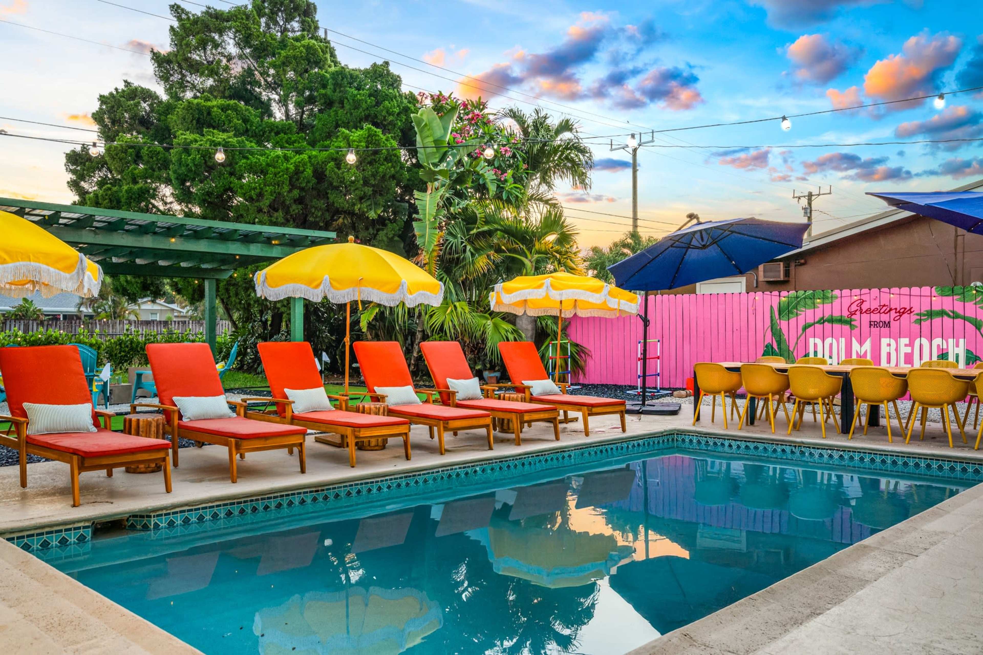 A pool area features lounge chairs with orange cushions, yellow umbrellas, and a pink mural, set against a backdrop of lush greenery and a sunset sky.