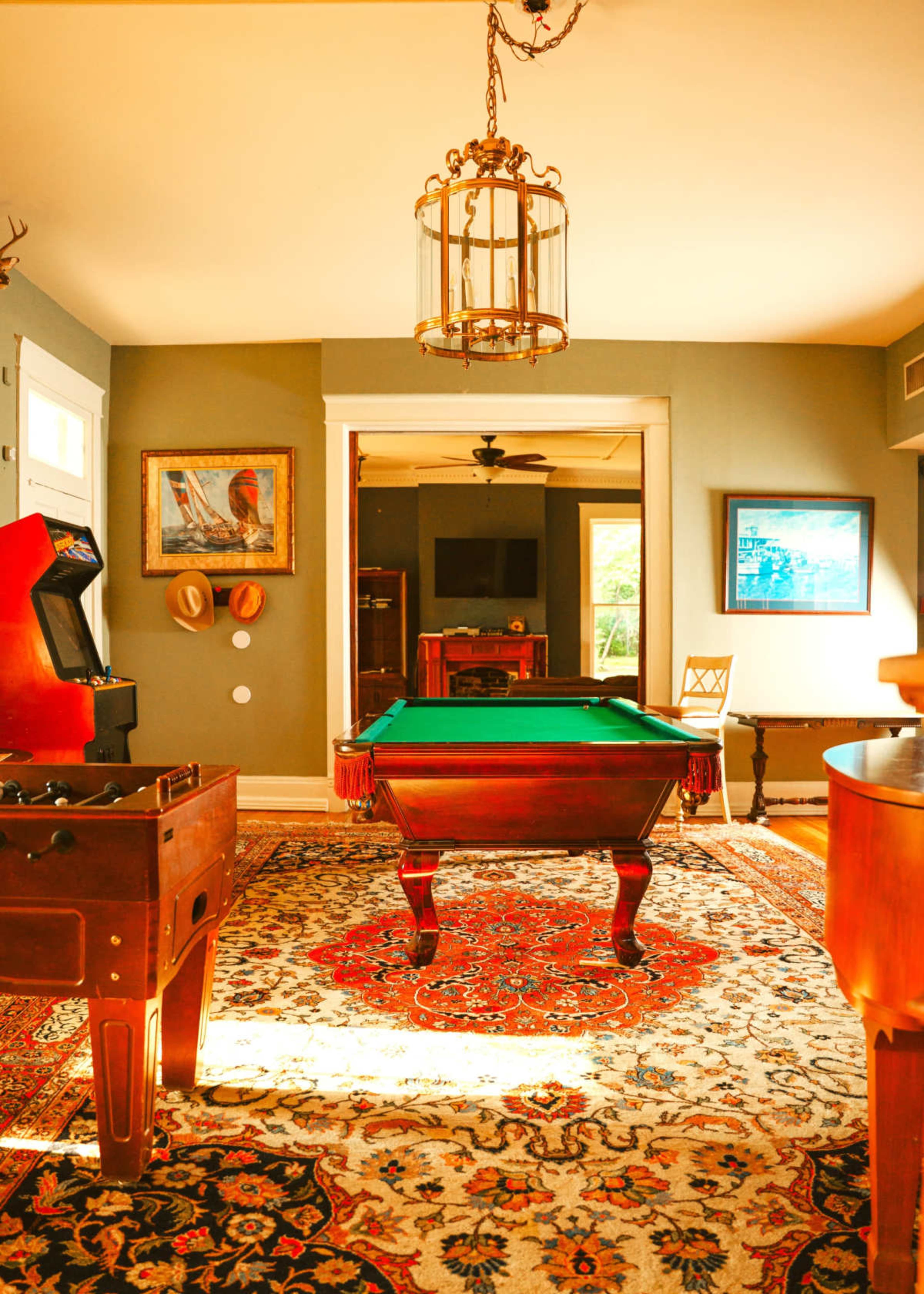 The image depicts a room featuring a billiard table, vintage arcade machine, and a decorative rug, with a warm light illuminating the space.
