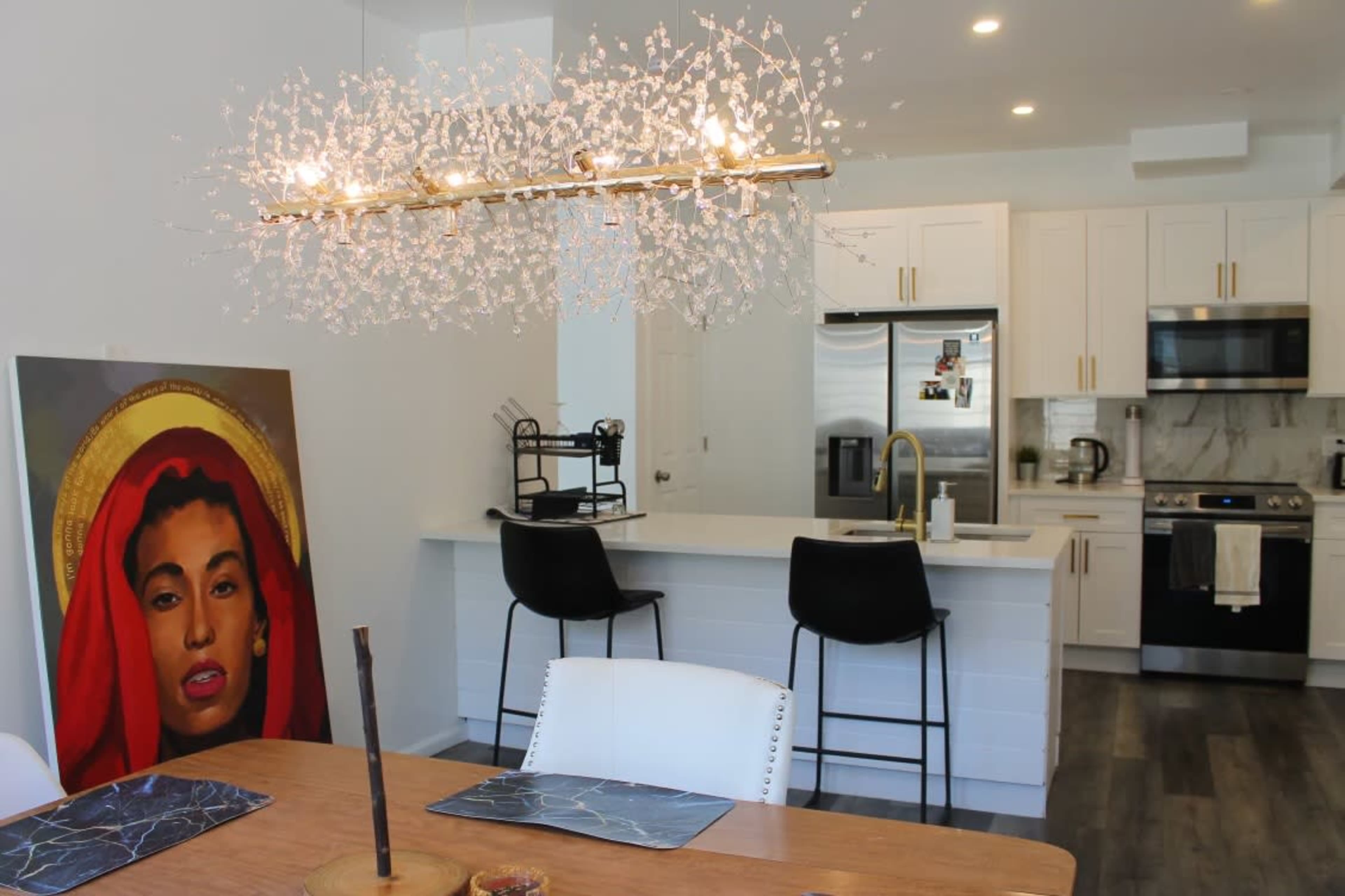 A modern kitchen features a central island with bar stools and a decorative chandelier, alongside a large painting of a woman with a red headscarf.