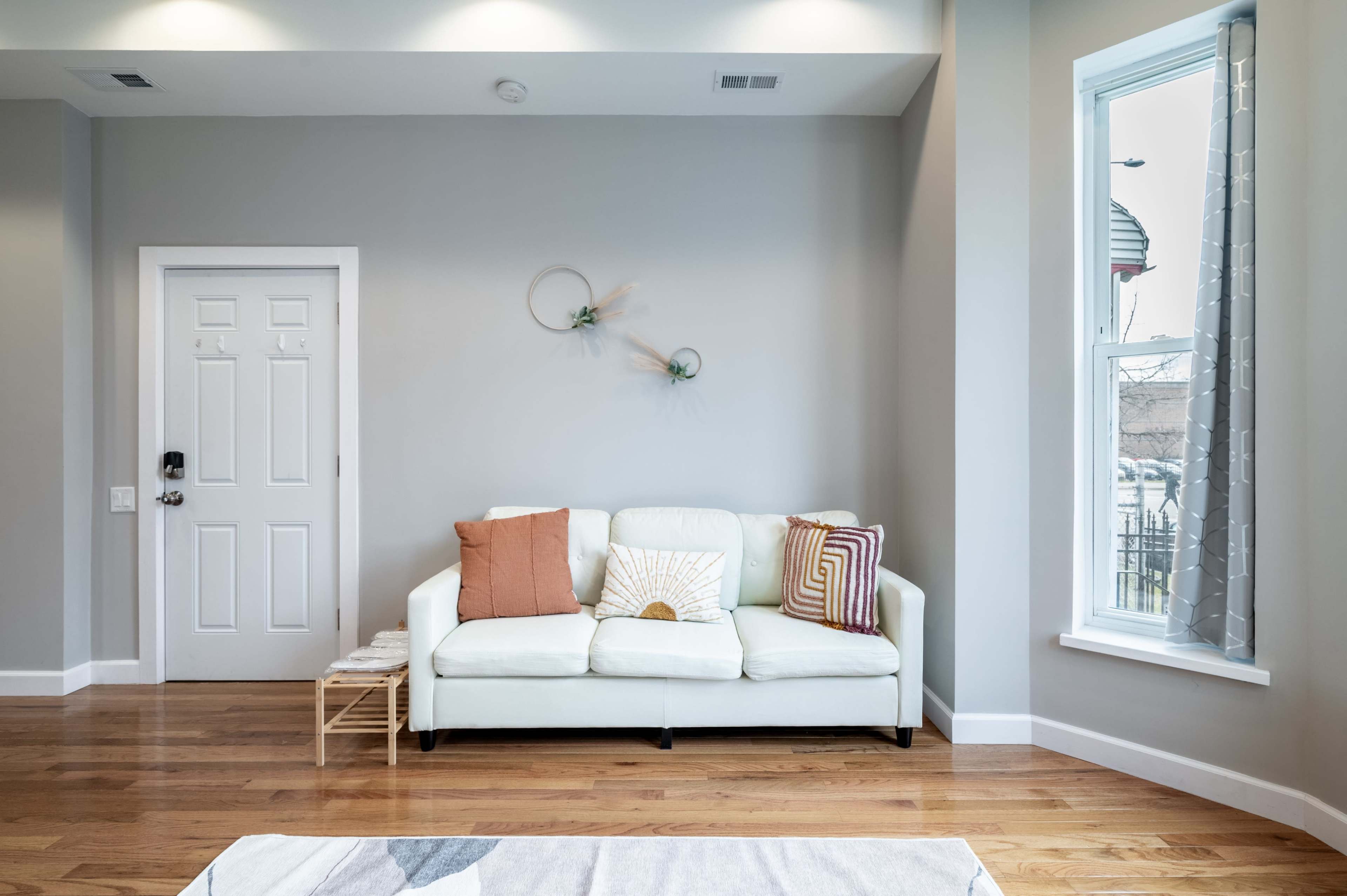 A white sofa with decorative pillows is situated against a gray wall in a room featuring a door and a large window.