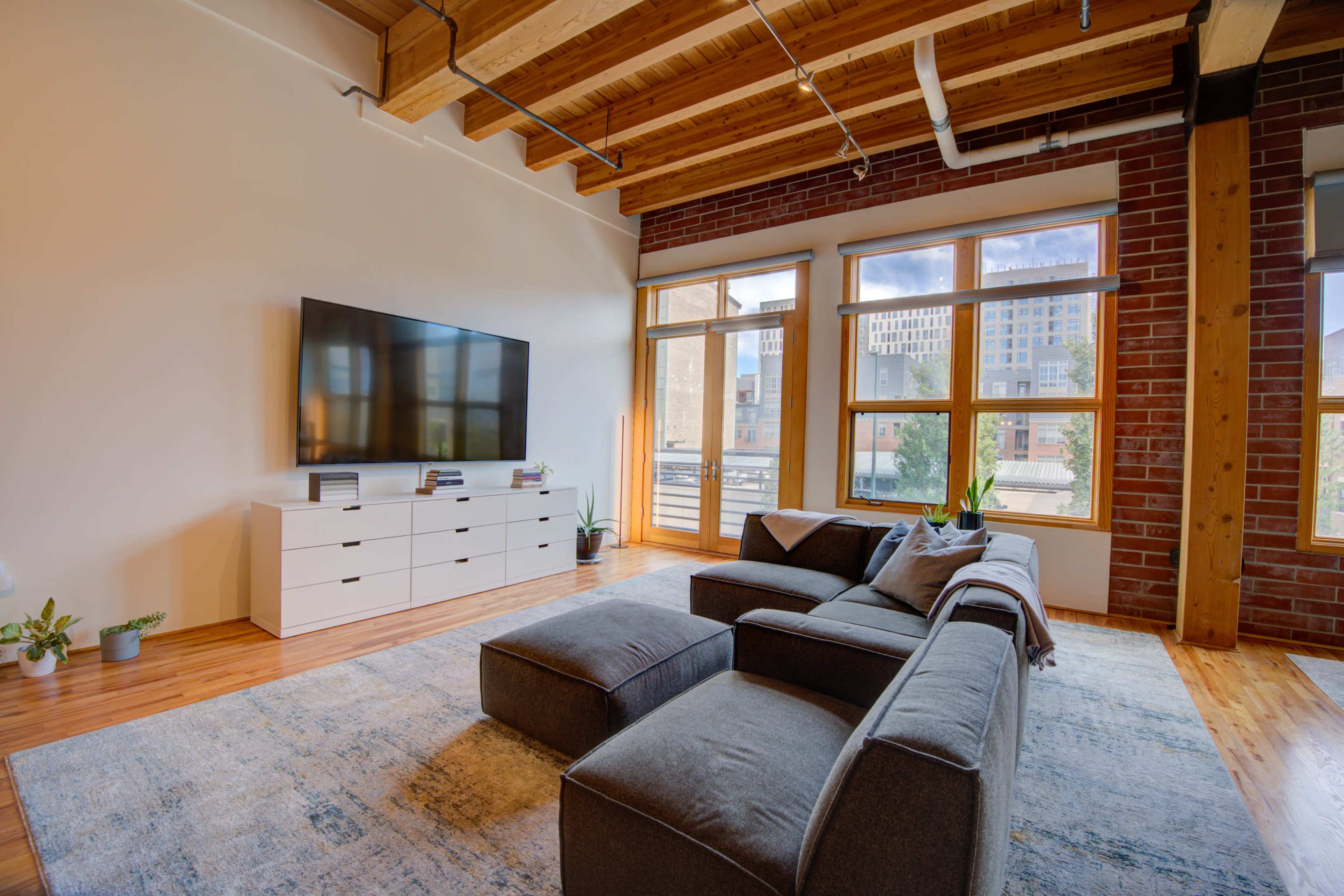 The image shows a modern living room featuring a large gray sectional couch, a white entertainment unit with a flat-screen TV, and large windows allowing natural light to enter.
