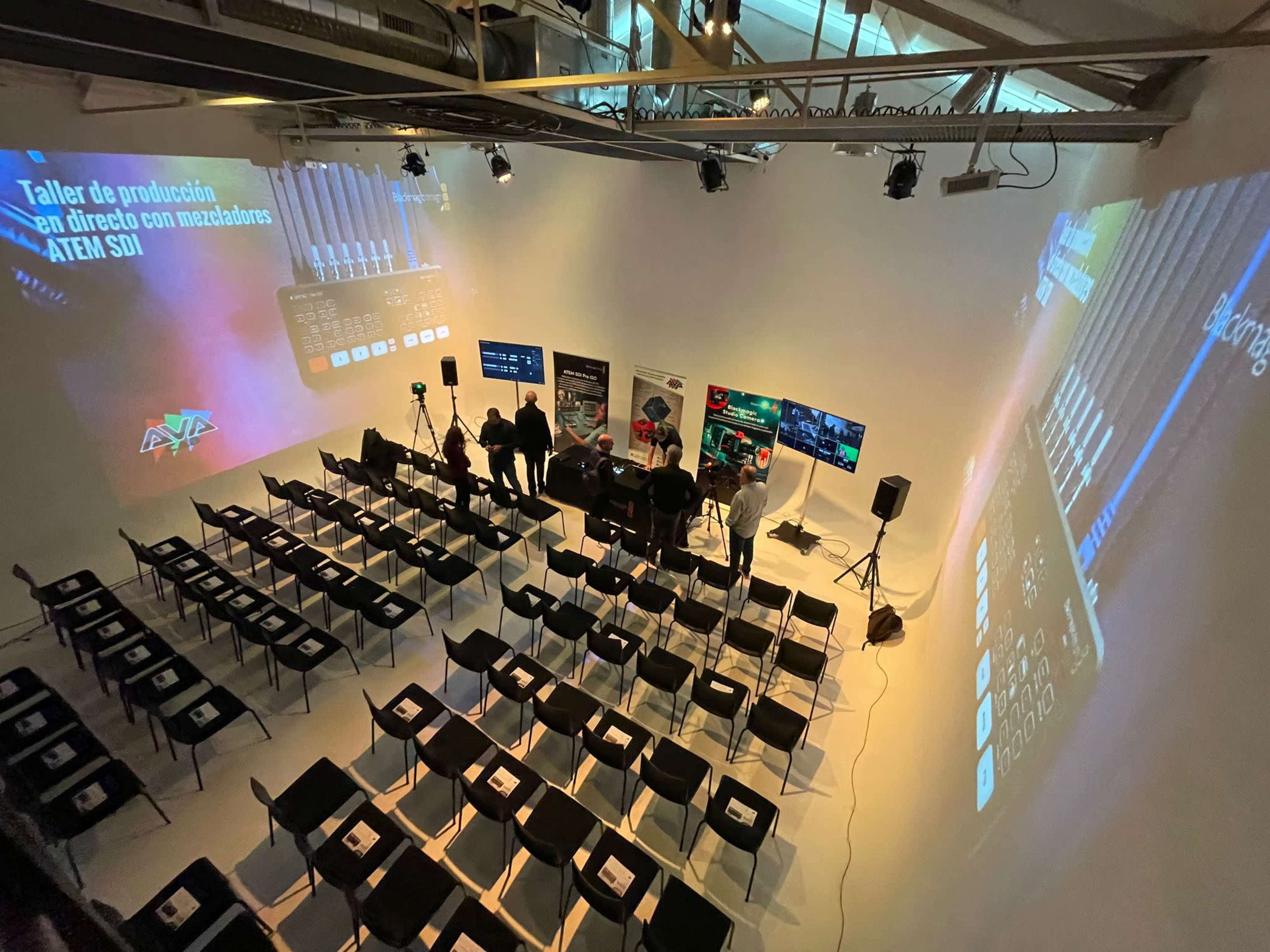 A spacious event space set up for a production workshop features rows of empty chairs and multiple screens displaying information about video production equipment.
