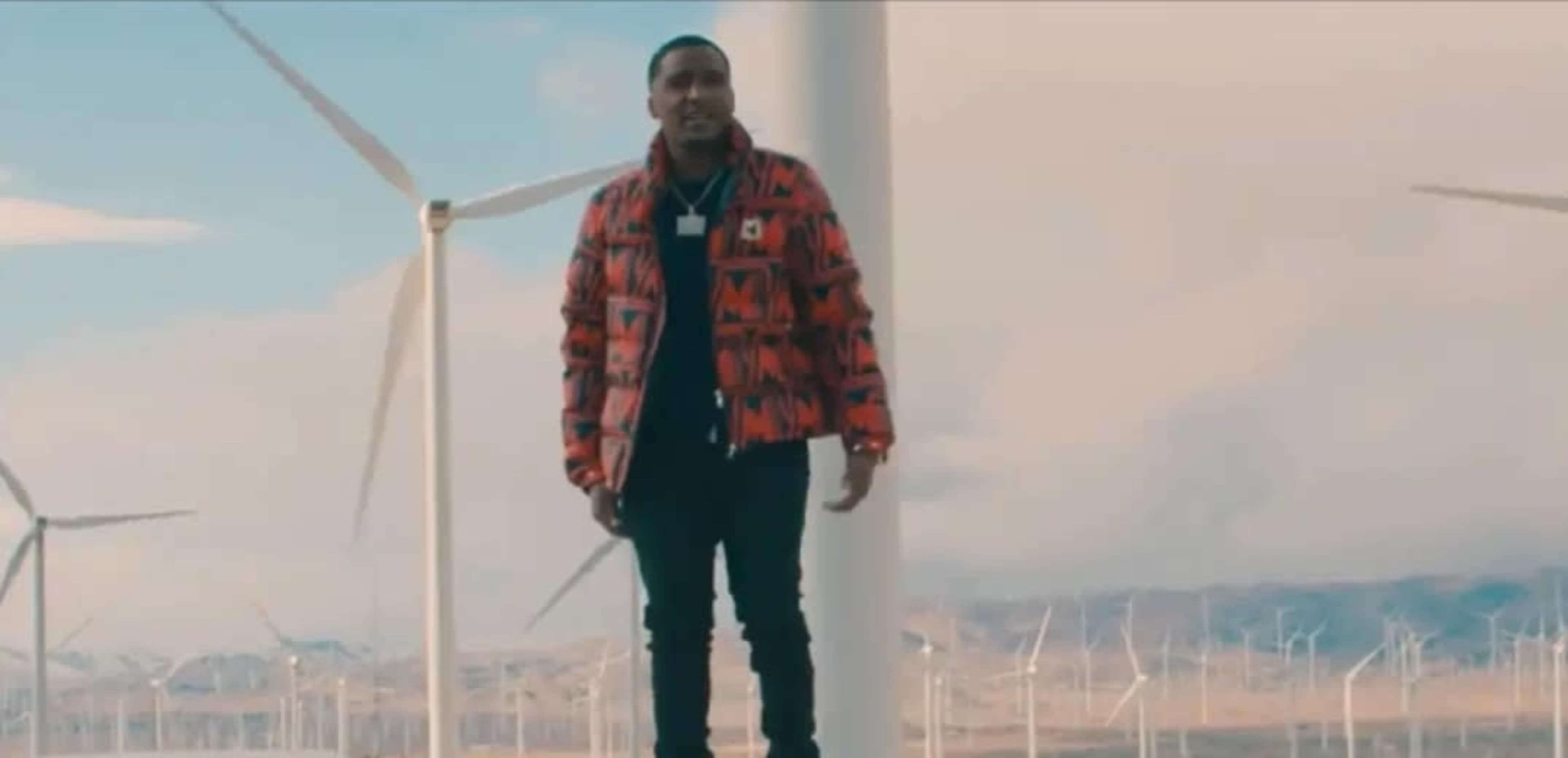 A man stands among large wind turbines against a backdrop of mountains and a cloudy sky.
