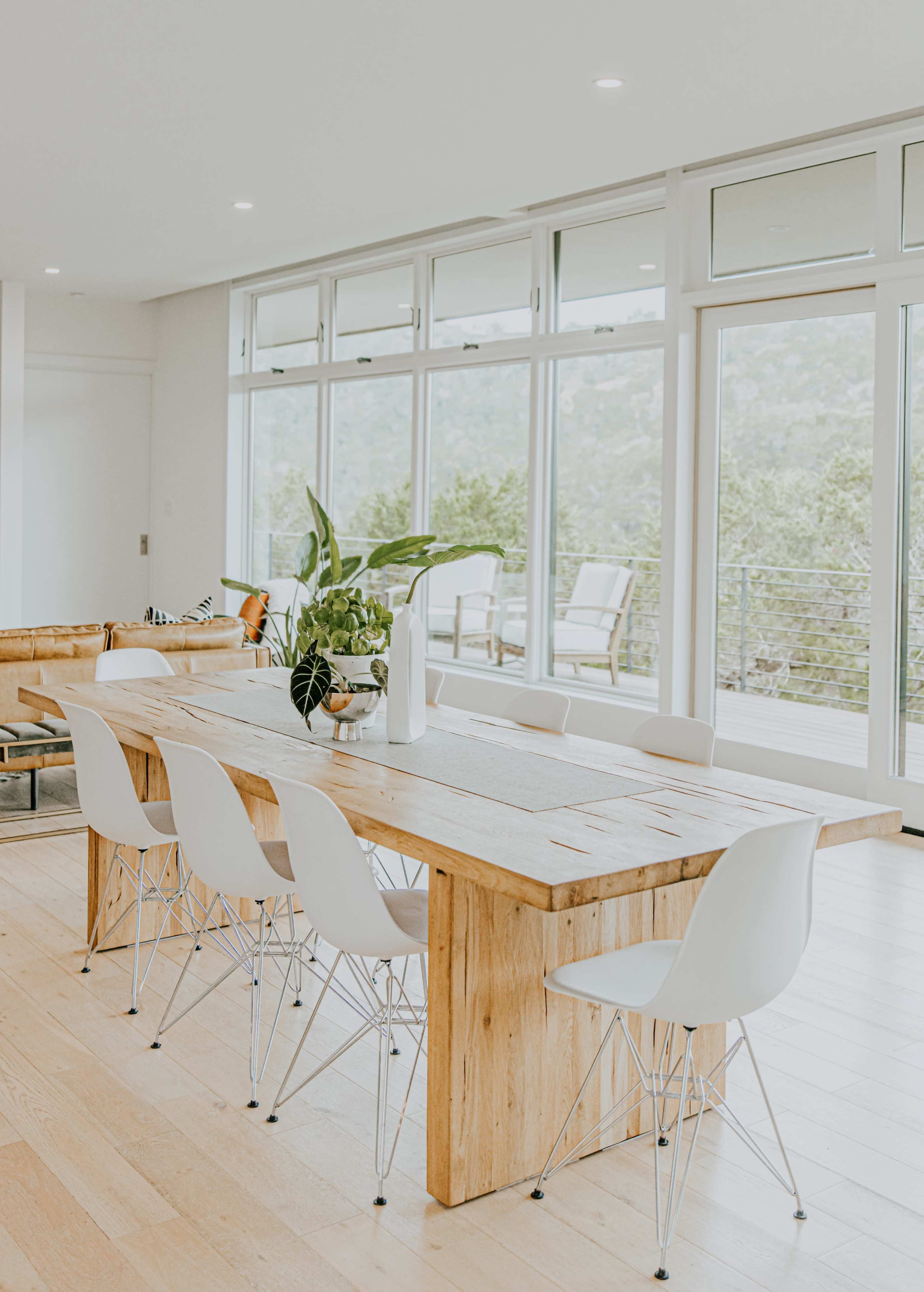 A wooden dining table with white chairs is set beside large windows that overlook a green landscape.