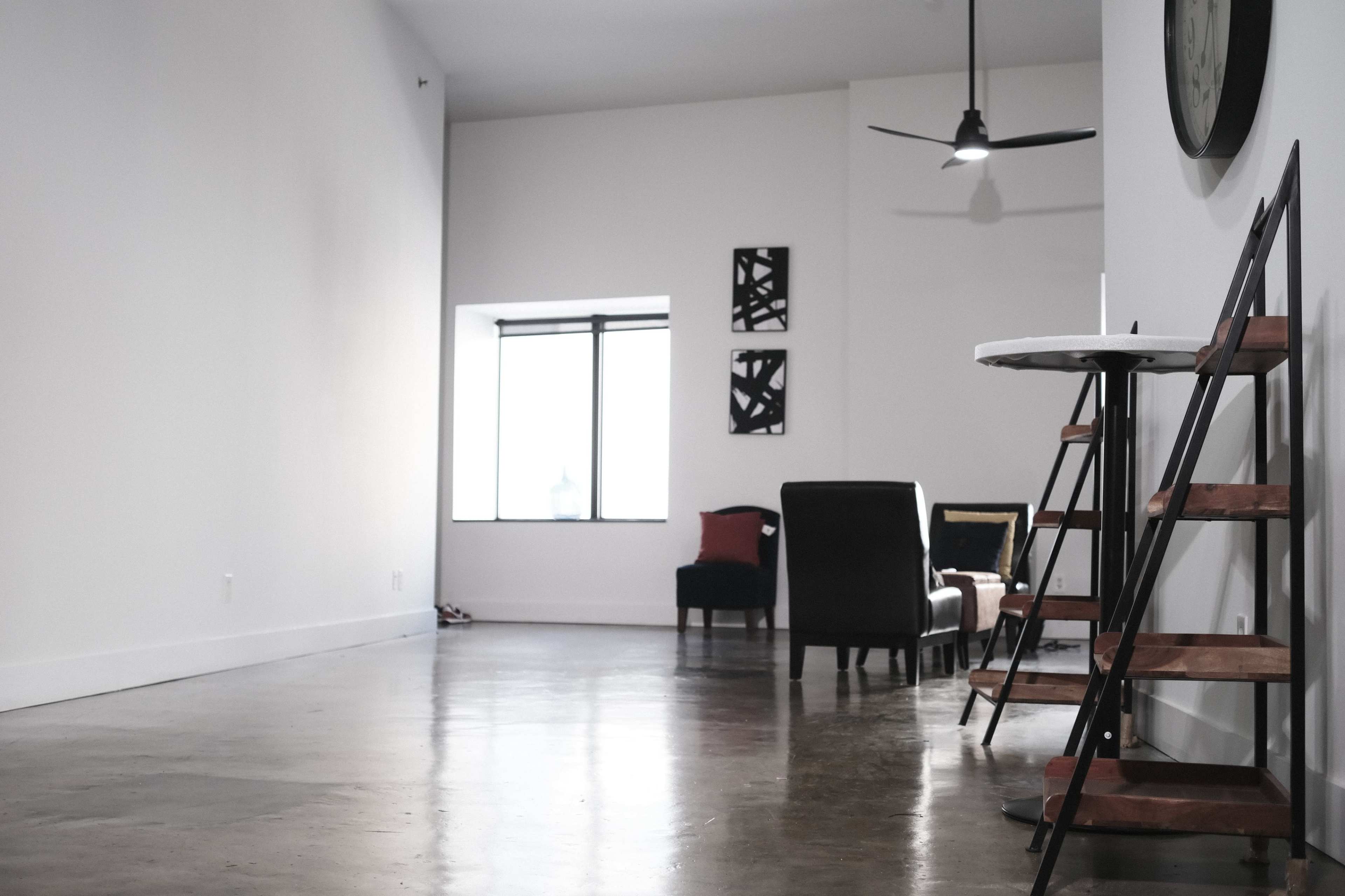 A minimalist interior space featuring a polished concrete floor, a large window, a seating area with a chair and side table, and a wooden ladder shelf against the wall.