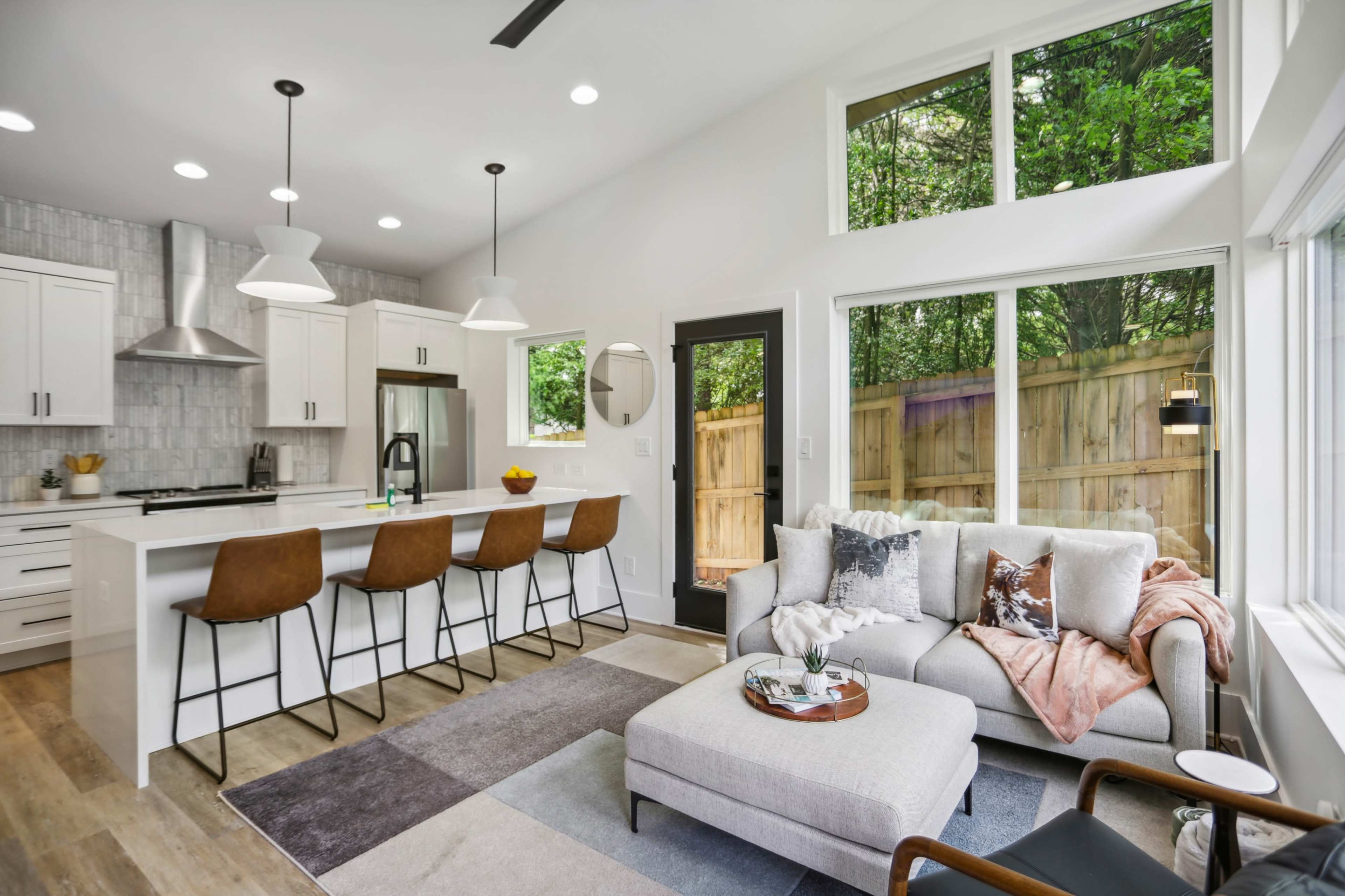 A modern kitchen and living area with a sleek design, featuring a large window overlooking a fenced yard, a gray sofa, and a central island with bar stools.