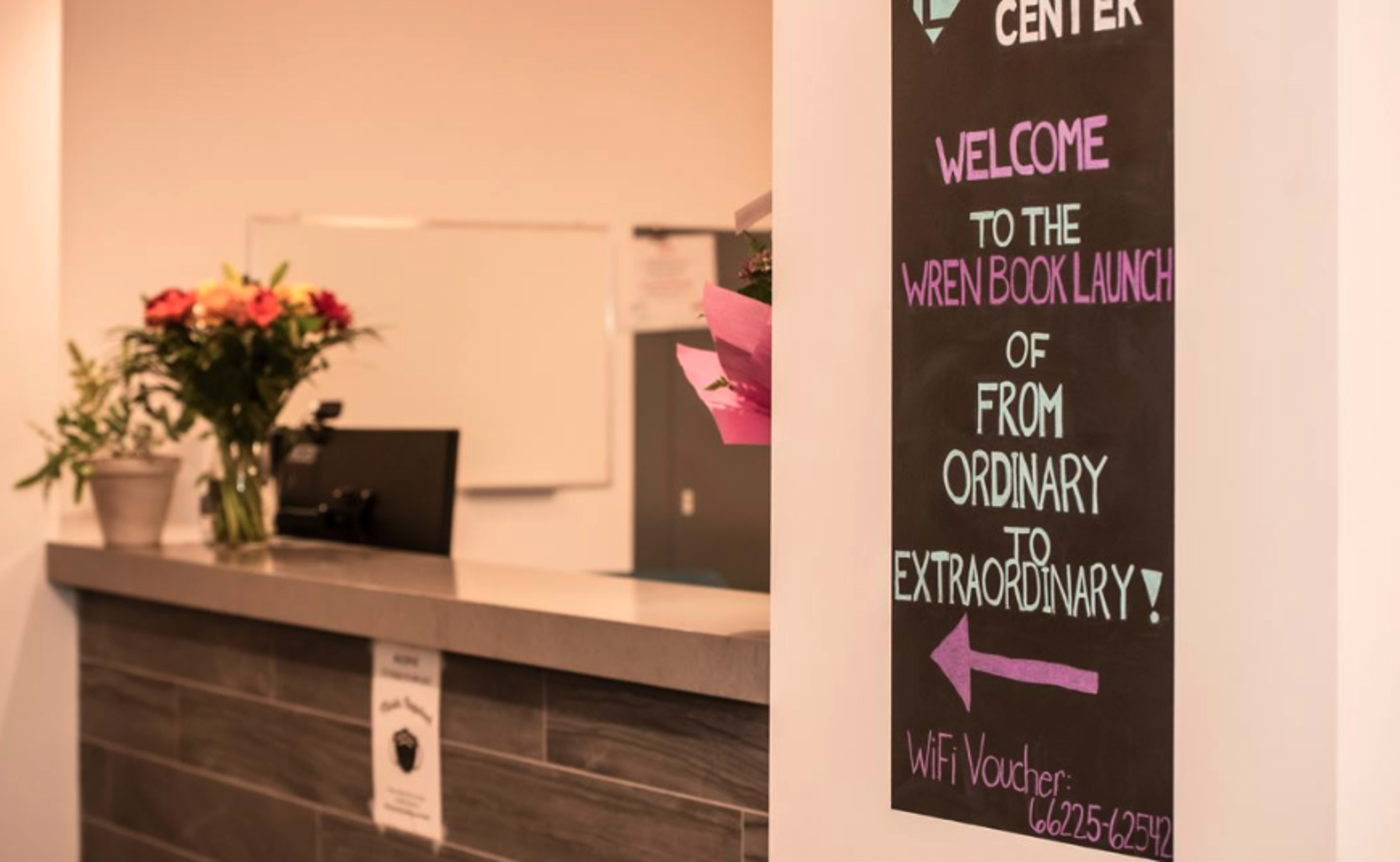 A welcome sign for the "Wren Book Launch" event at a reception desk, with flowers and a WiFi voucher number displayed.