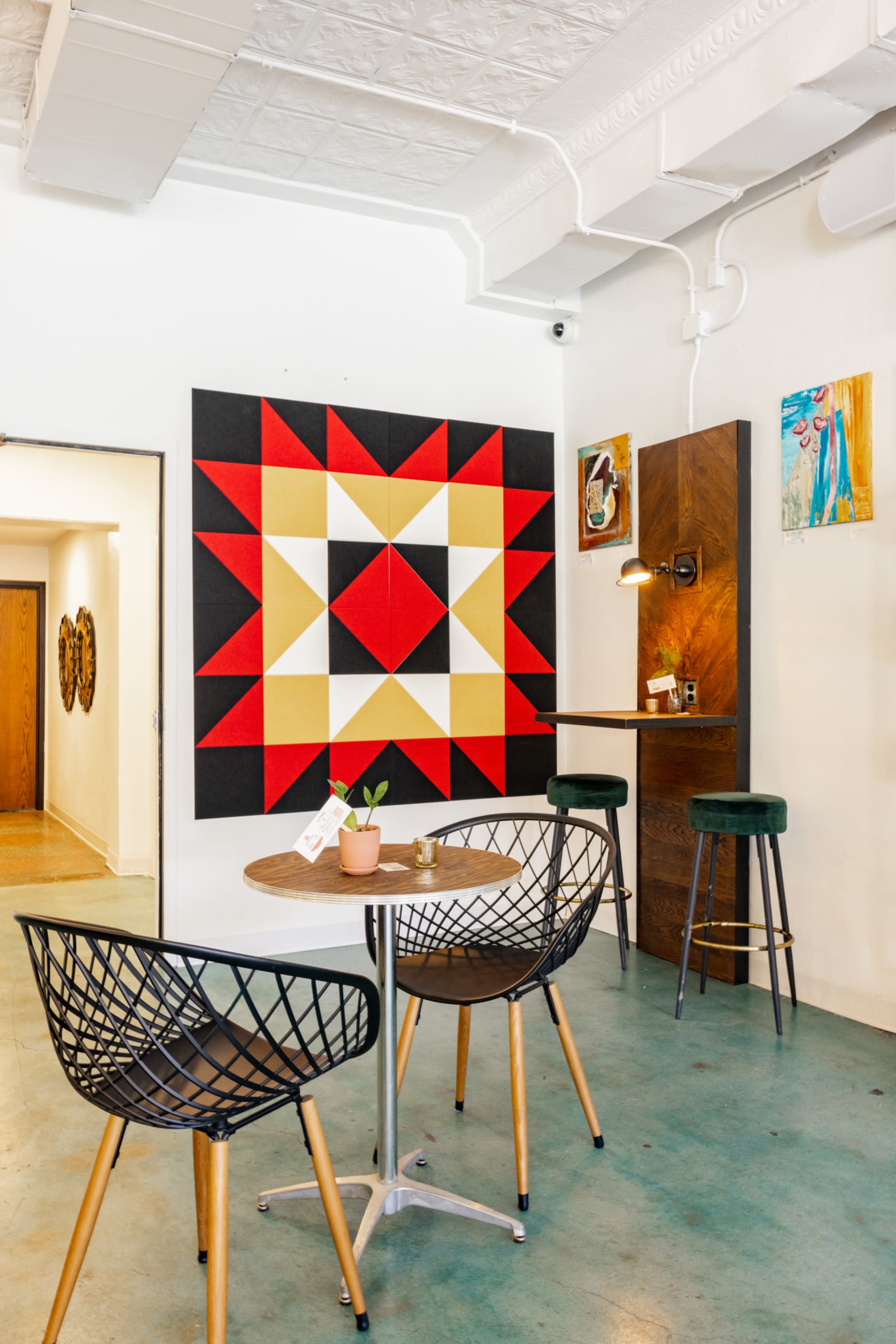 A modern café interior featuring a geometric wall art piece in red, black, and yellow, with a small table and chairs arranged beside a wooden bar.