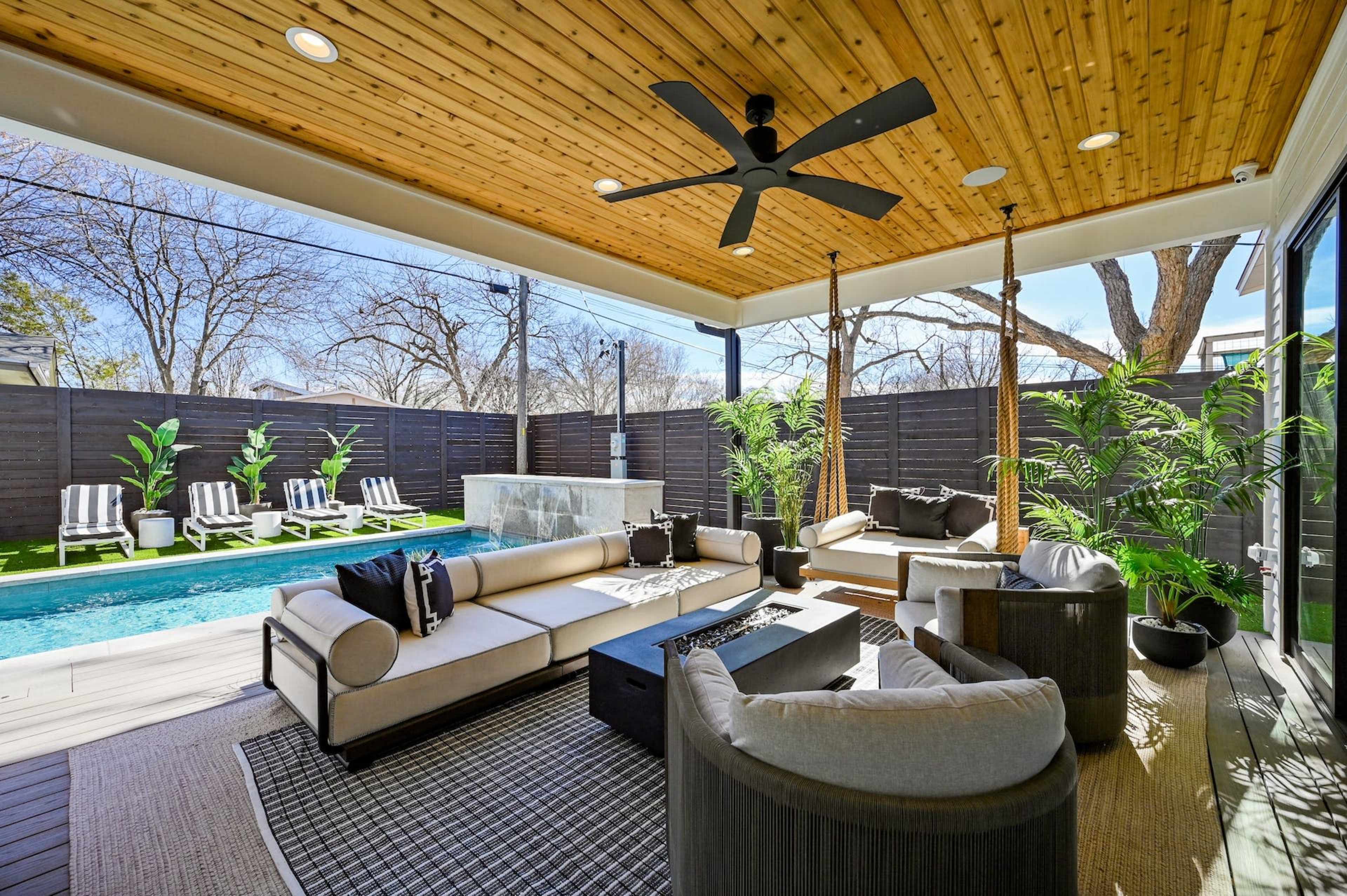 The image shows a covered outdoor patio with a seating area, a swimming pool in the background, and trees visible beyond a privacy fence.