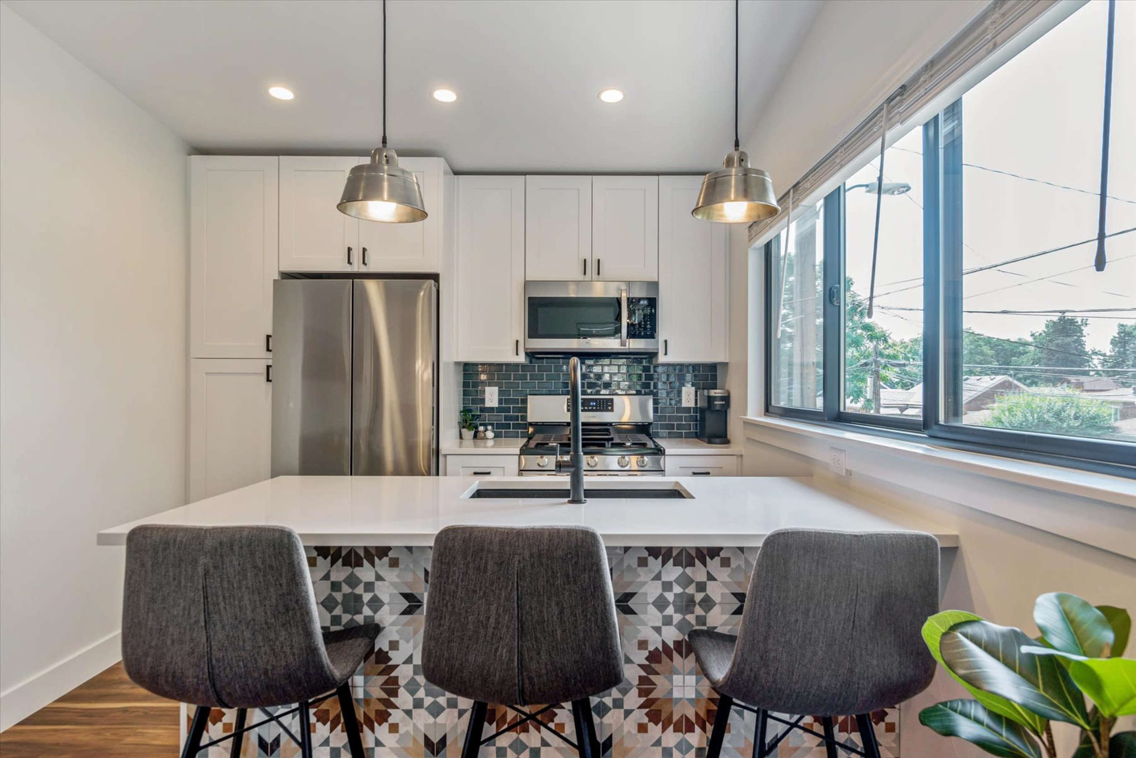 A modern kitchen features a large island with three stools, stainless steel appliances, and stylish pendant lights.