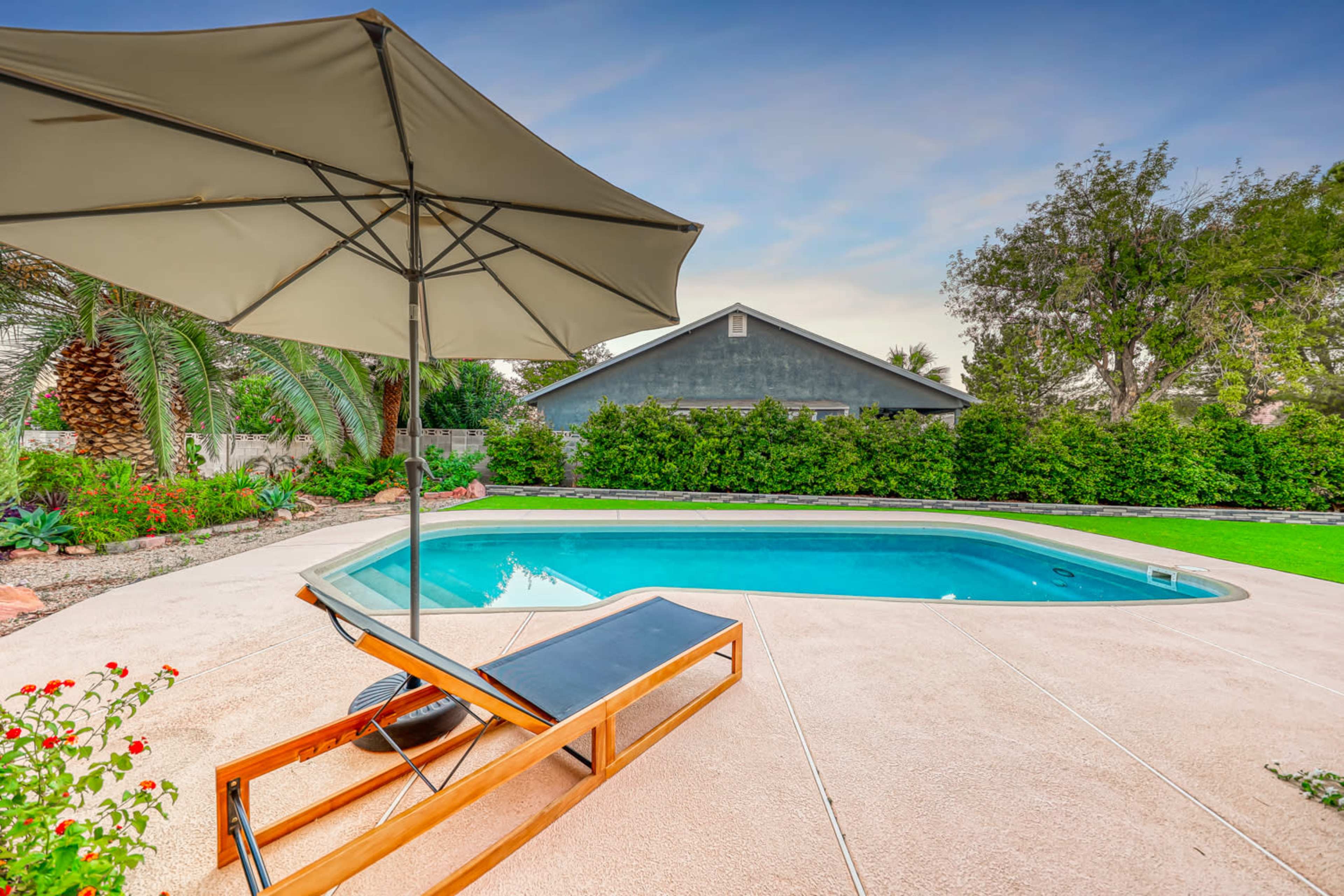 A patio with a lounge chair and umbrella overlooks a swimming pool surrounded by greenery and a house in the background.
