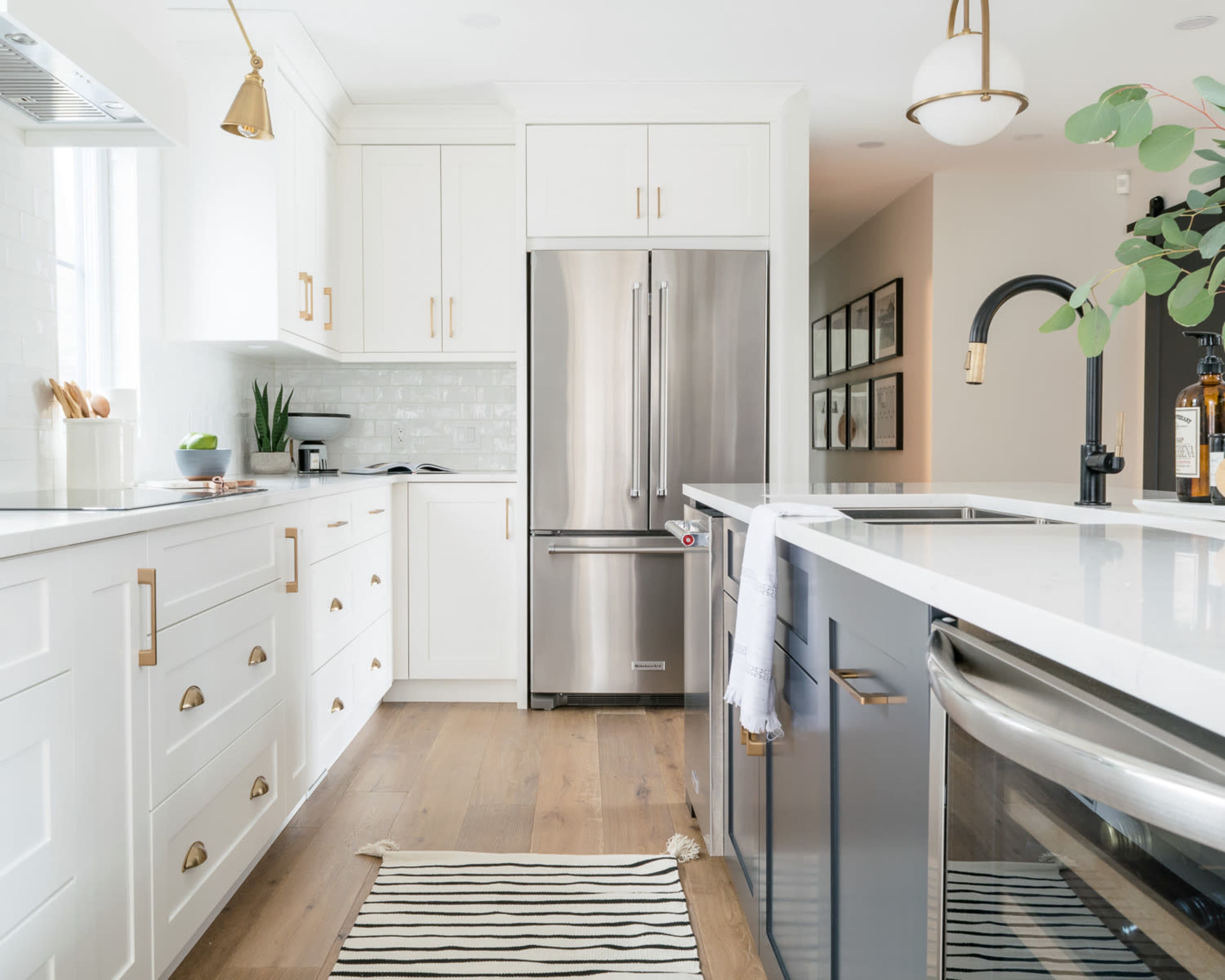 The image shows a modern kitchen with white cabinetry, stainless steel appliances, and a striped rug on a wooden floor.