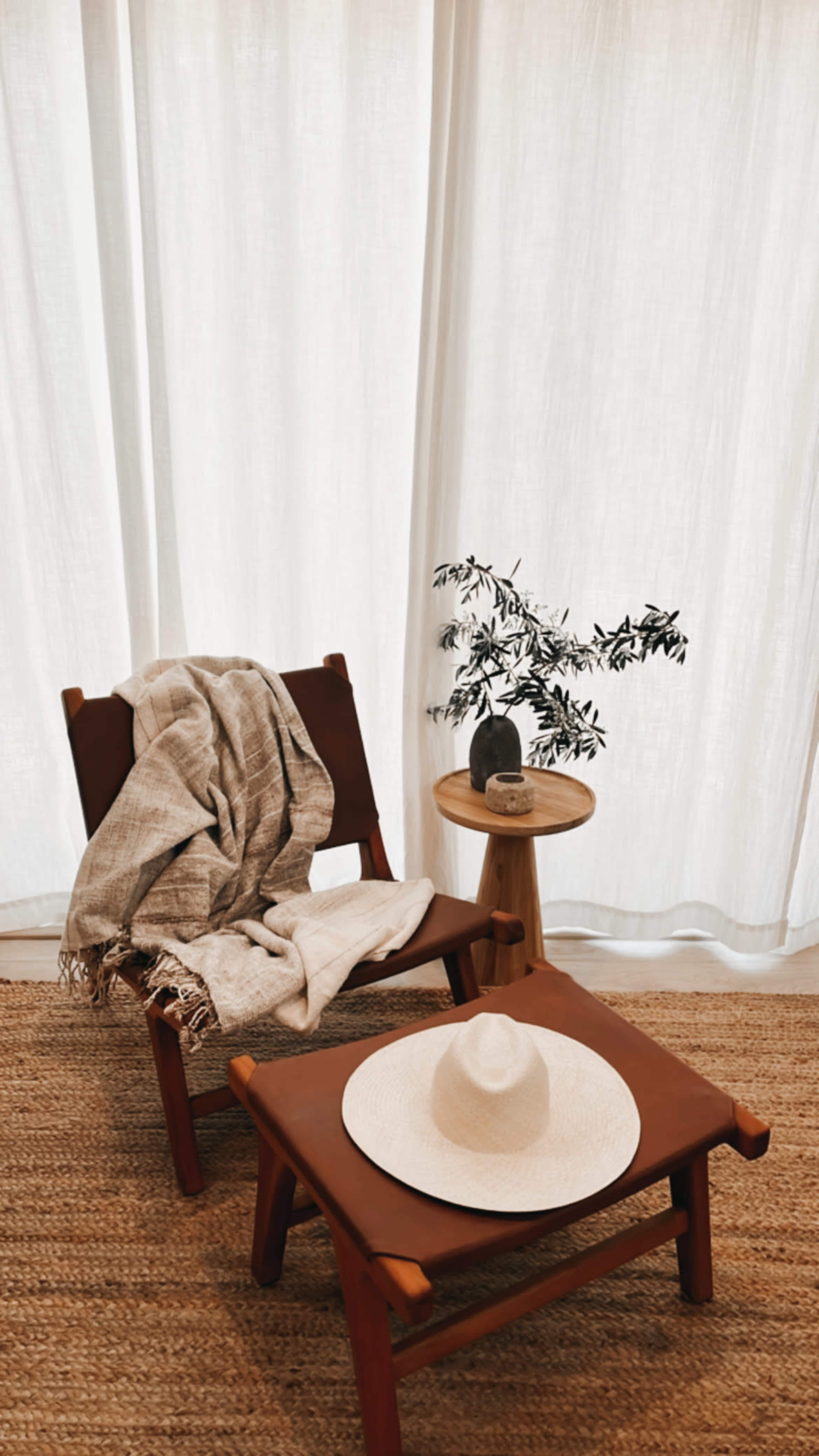 A wooden chair with a blanket draped over it sits beside a small round table, which holds a decorative object and a potted plant, all set against a backdrop of sheer white curtains.