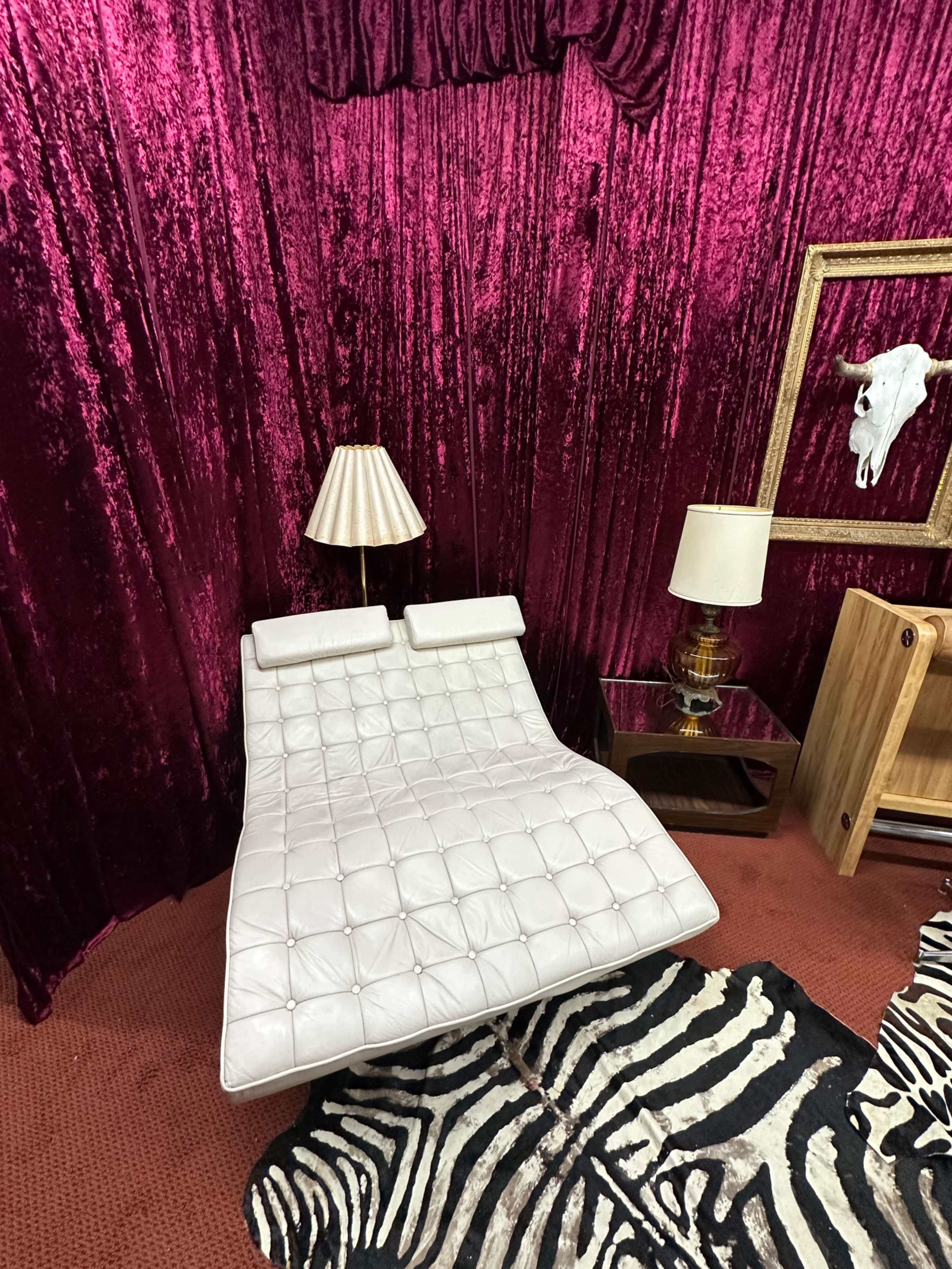 The image shows a modern white chaise lounge in a room with shiny magenta curtains, accompanied by a lamp and a zebra-striped rug.