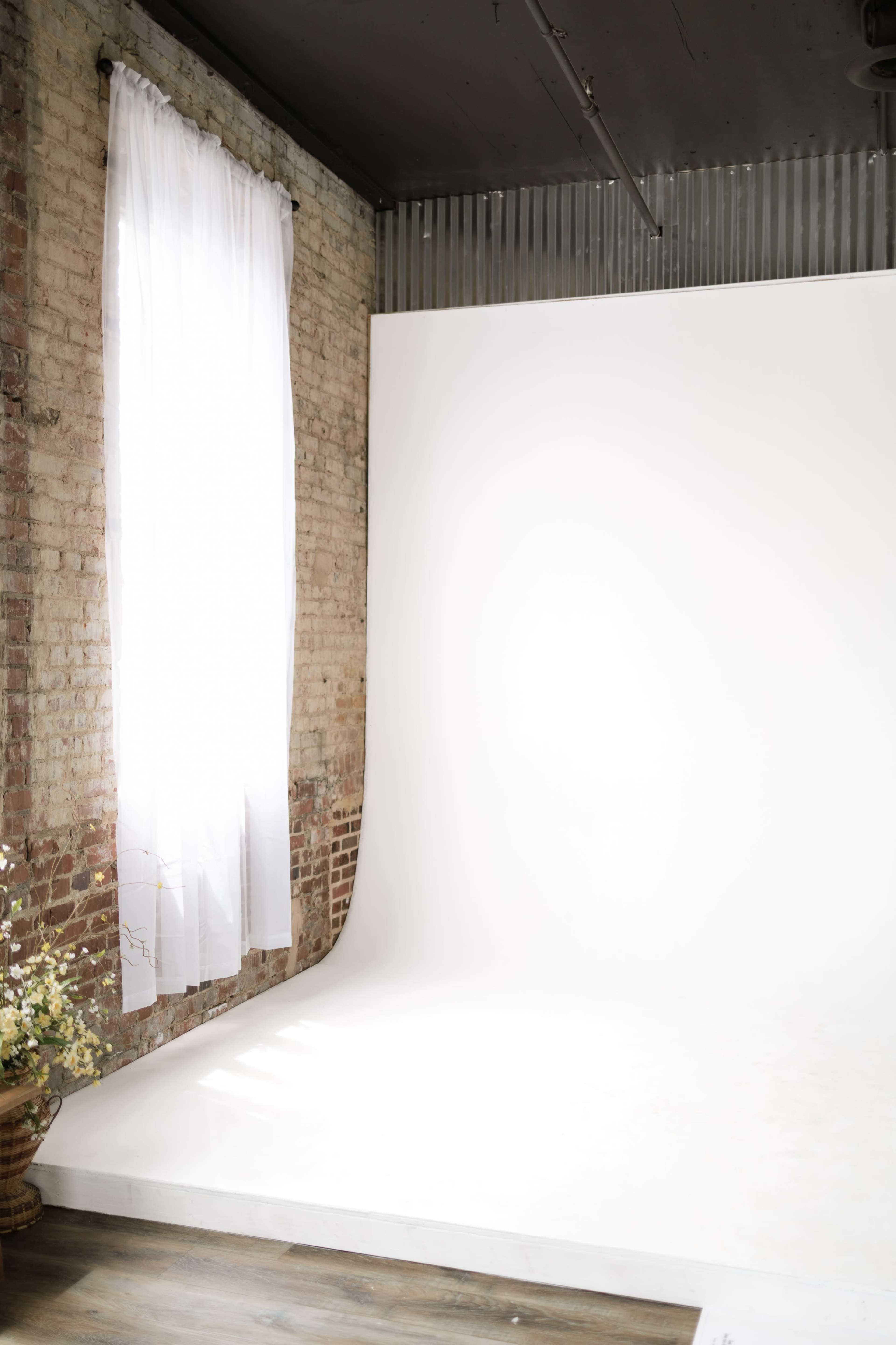 A photography studio features a white backdrop and a large window with sheer curtains, allowing natural light to illuminate the space.