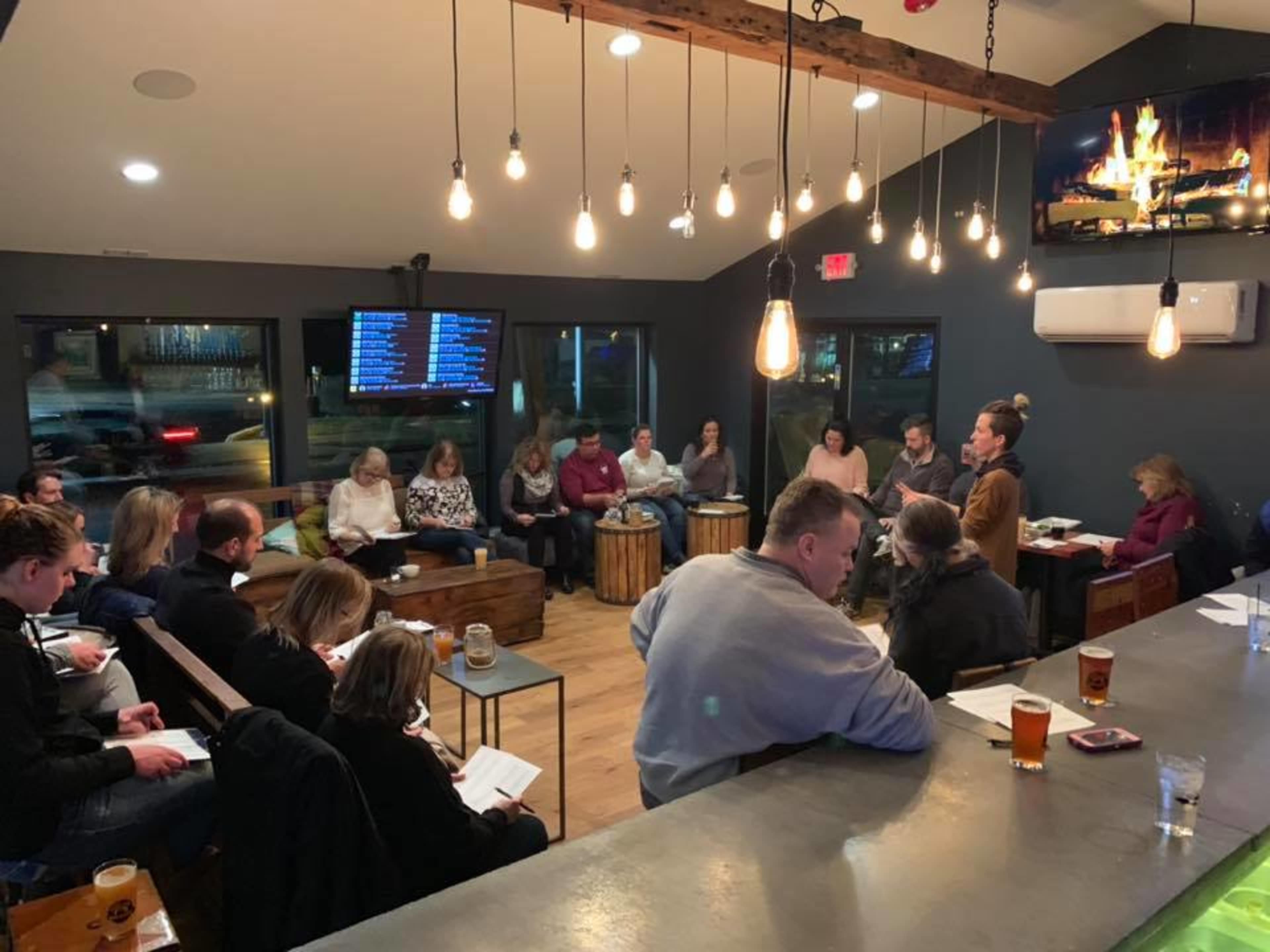 A group of people sits in a well-lit, modern café setting, engaging in discussion around a central speaker while others take notes.