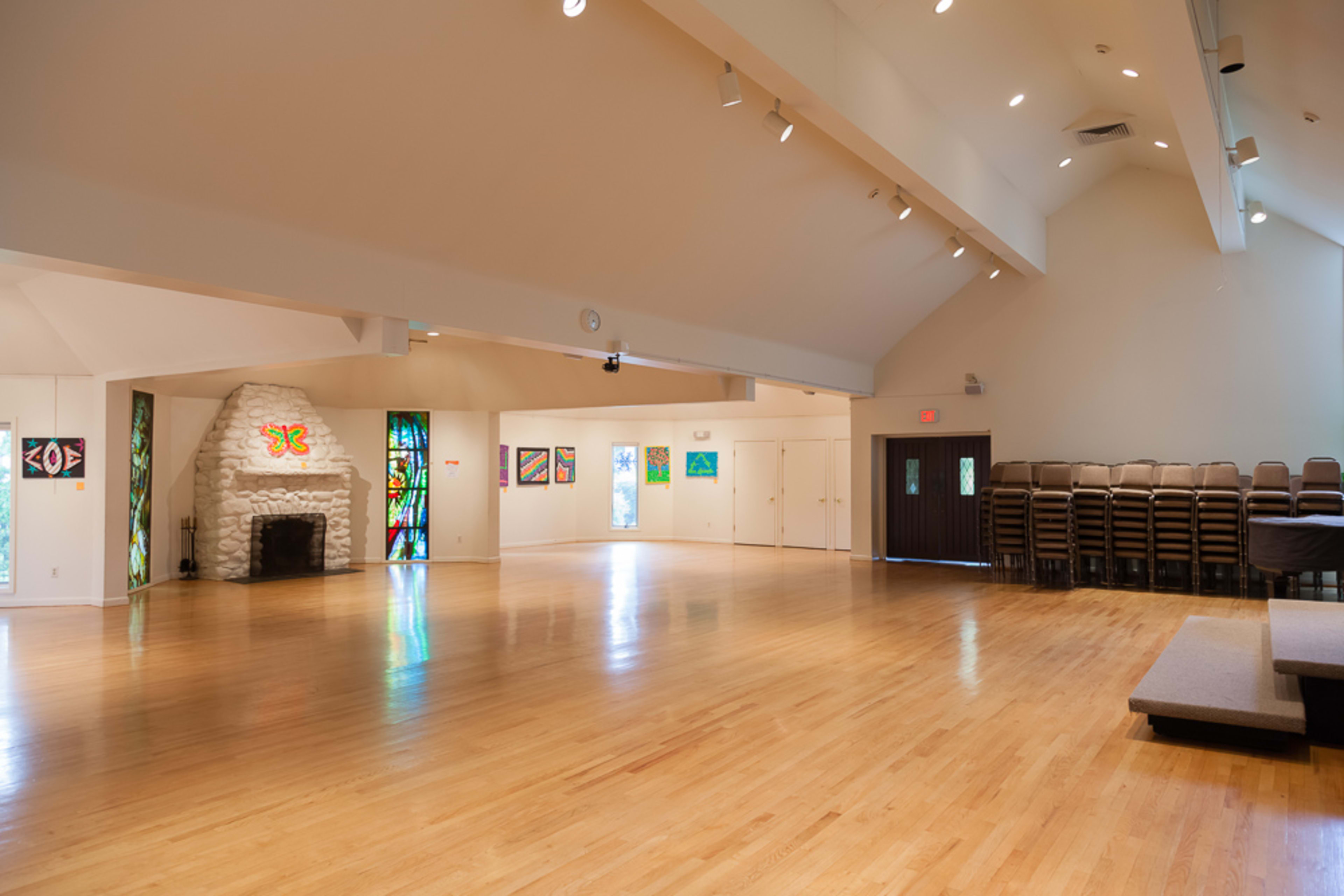 The image shows a spacious, brightly lit interior of a hall featuring wooden floors, a stone fireplace, and colorful stained glass windows.