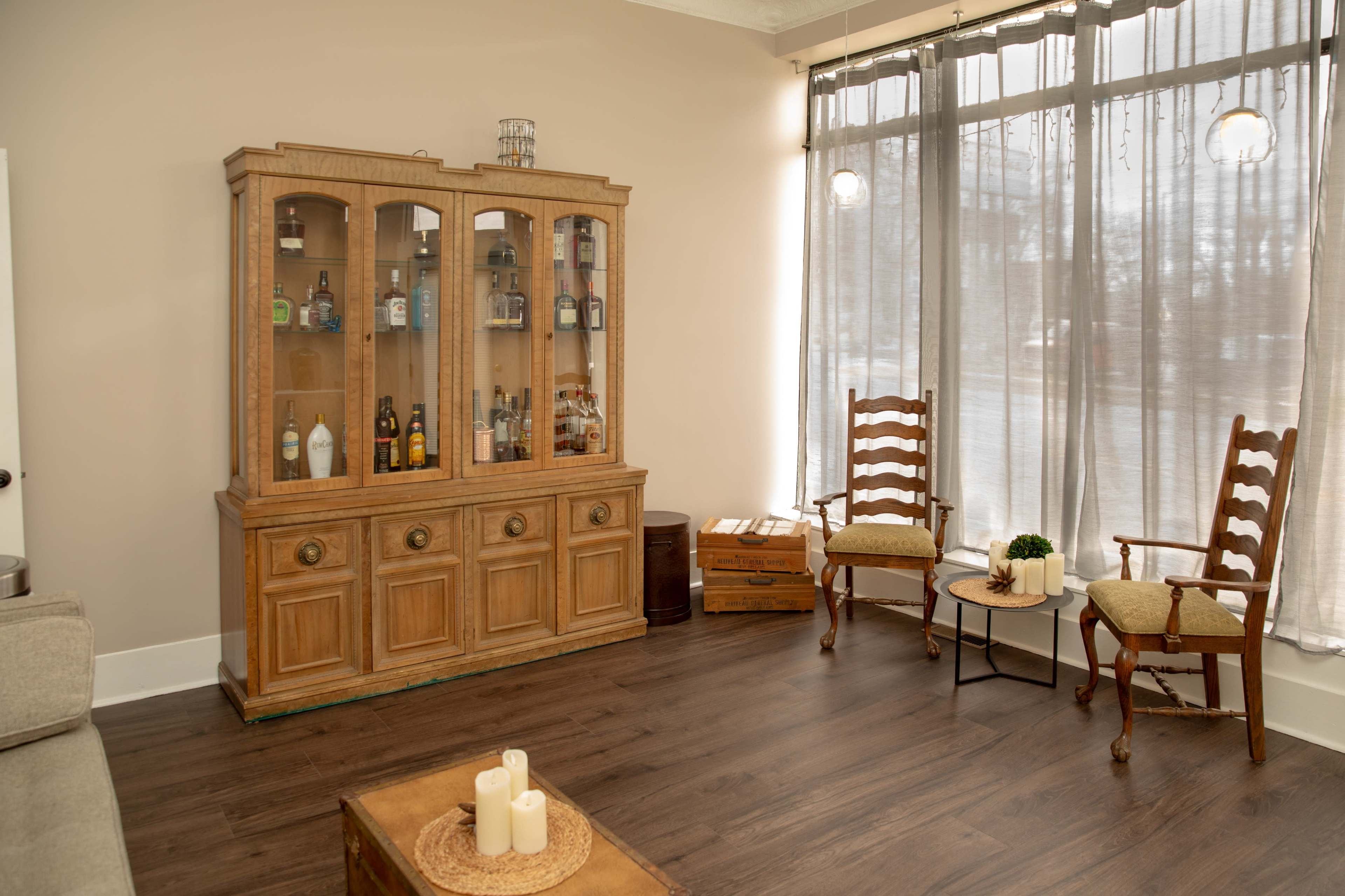 The image shows a living room with a light-colored wall, a wooden cabinet displaying various bottles, two wooden rocking chairs by a large window, and a small table with candles.