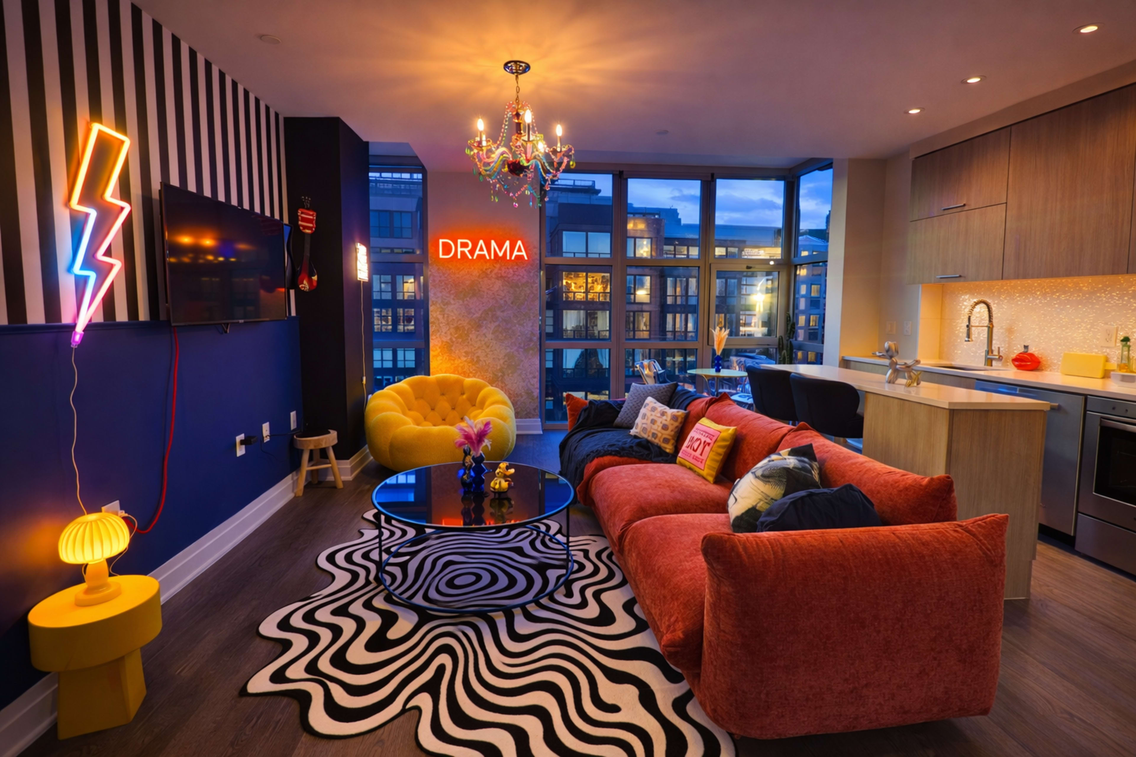 A modern living room features colorful furniture, a stylish rug, and a vibrant "DRAMA" sign, with large windows showcasing a cityscape at dusk.