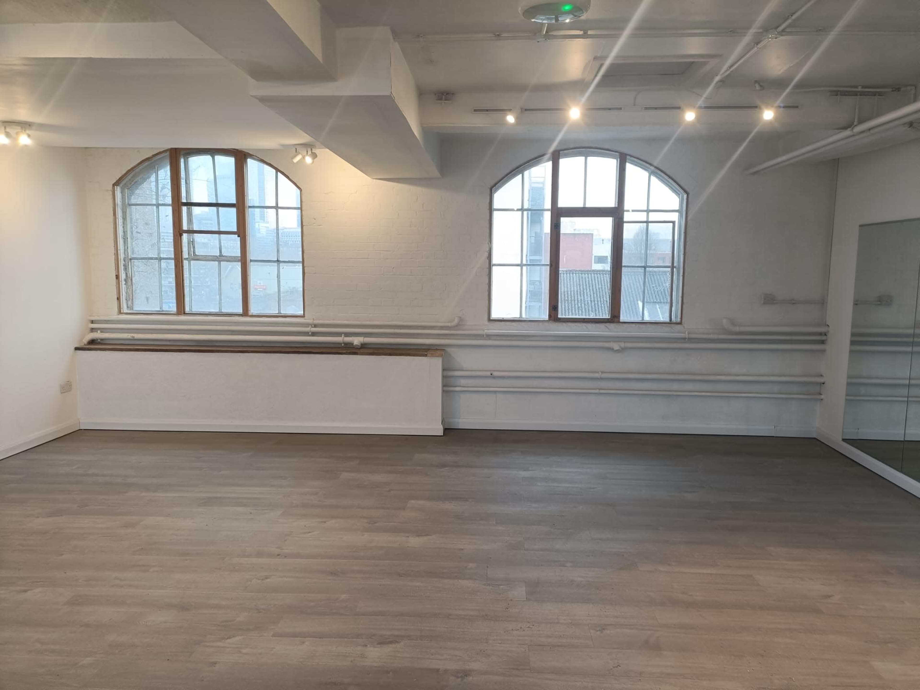 The image shows an empty, well-lit room featuring large windows, light wood flooring, and a mirror on one wall.