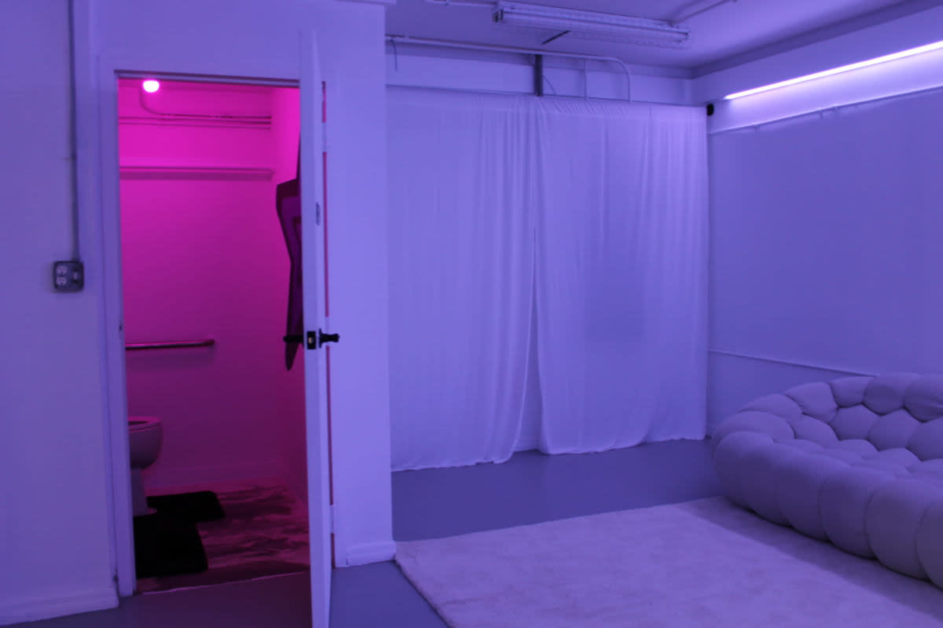 The image shows a white room with purple lighting, featuring a tufted couch, a doorway leading to a bathroom, and a curtain separating areas.