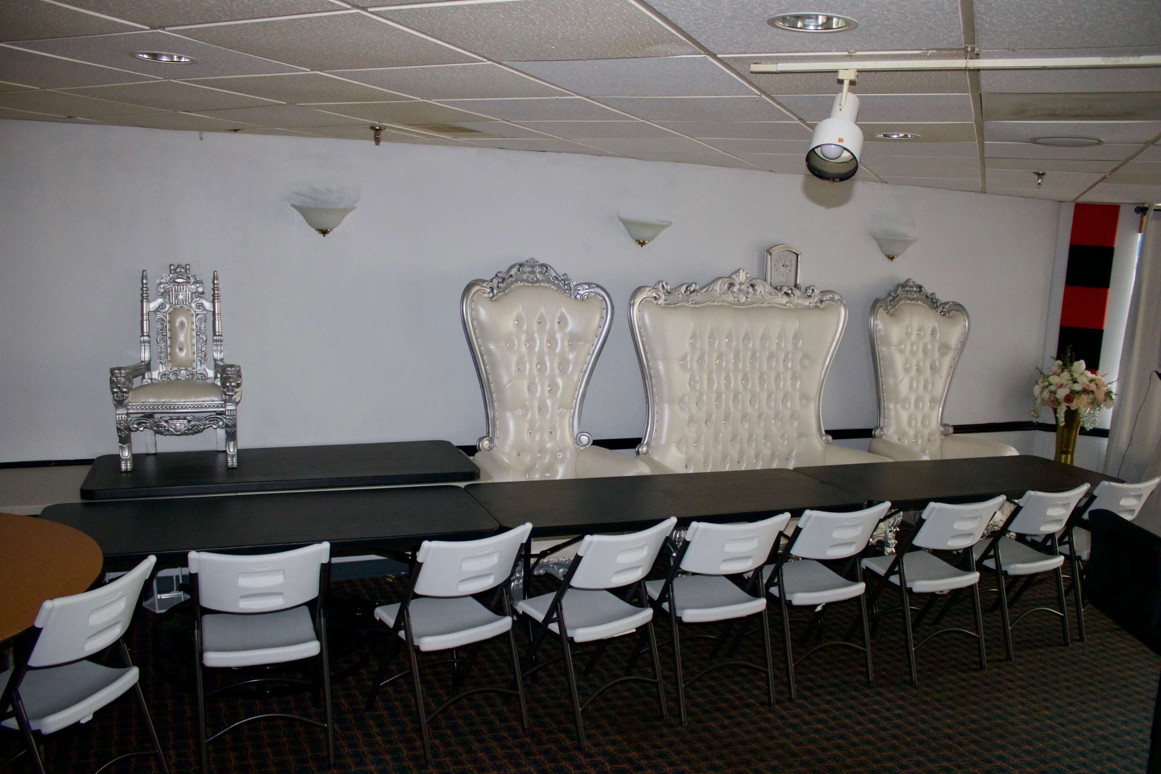 The image features a room set up for an event, with a long table and several white chairs arranged in front of three large, ornate throne-like chairs against a wall.