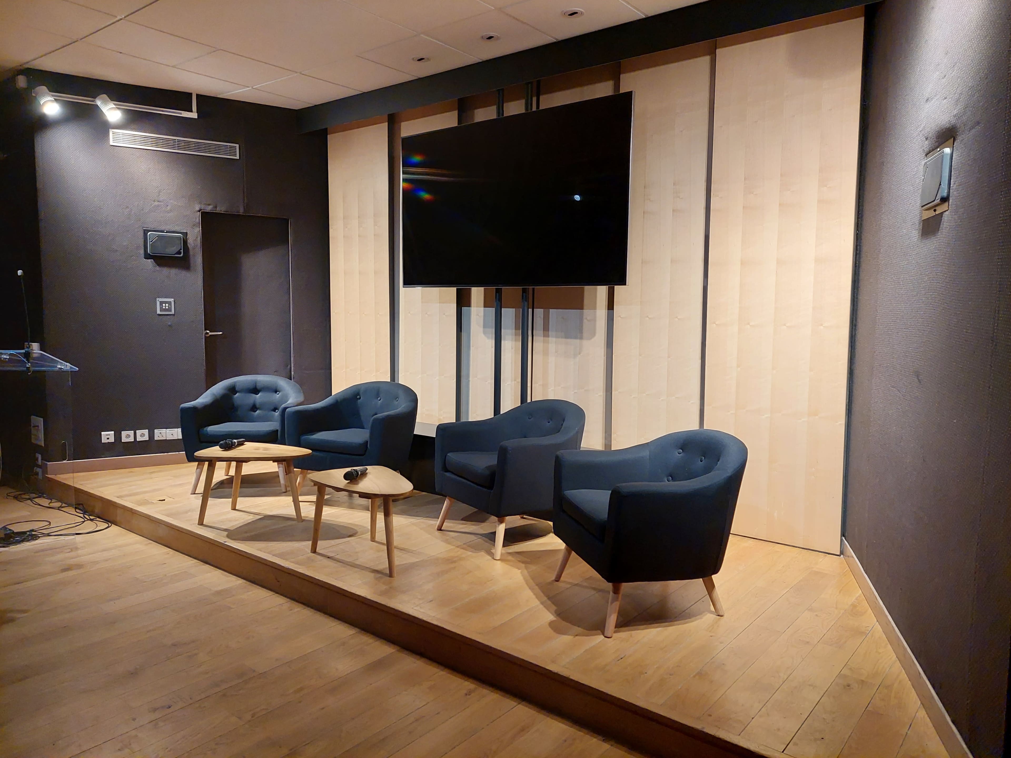 A conference room setup with three blue armchairs and a small table on a raised wooden platform, alongside a large wall-mounted screen.