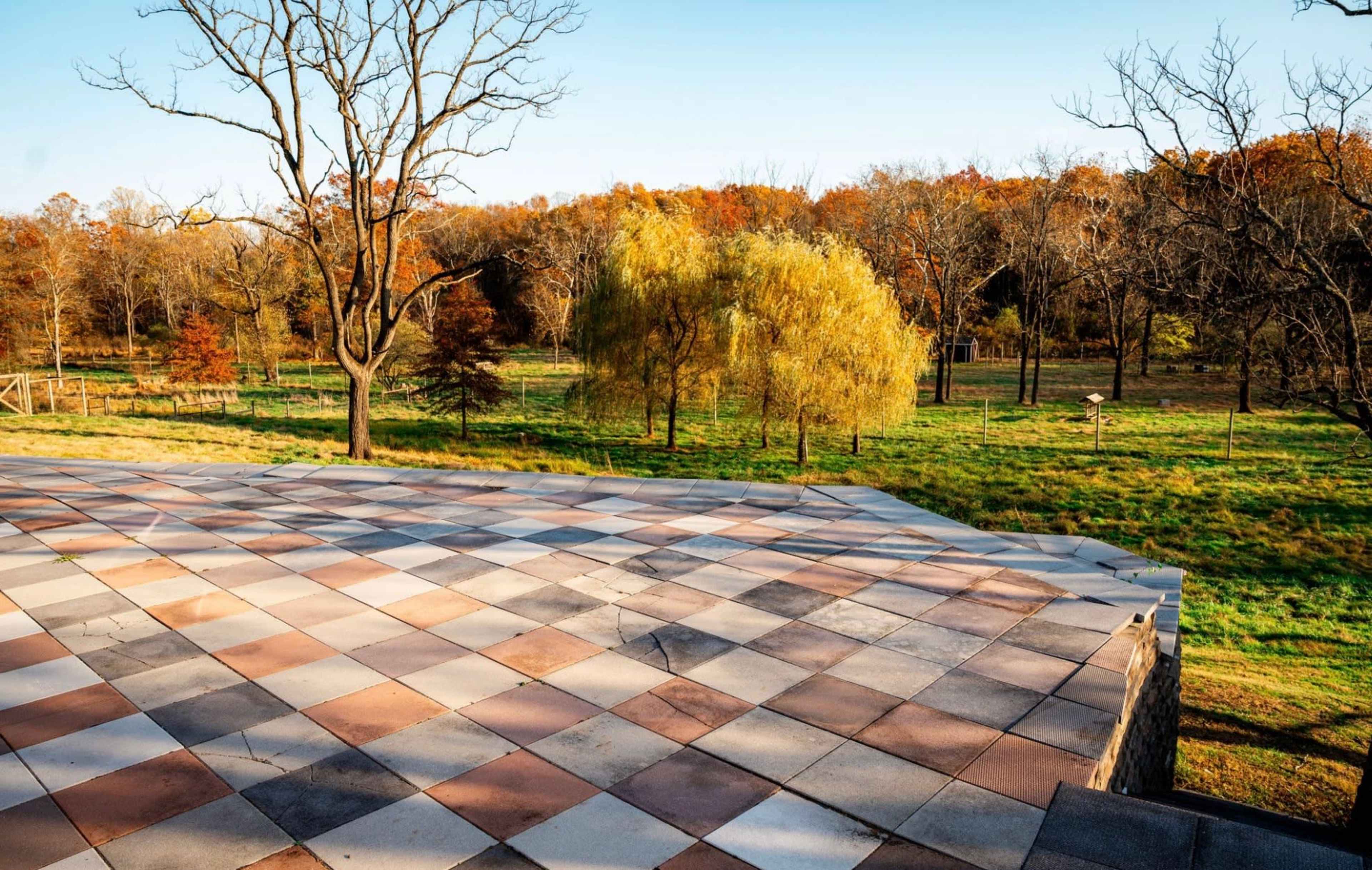 A sunlit patio with a checkerboard pattern overlooks a grassy field and trees in autumn foliage.