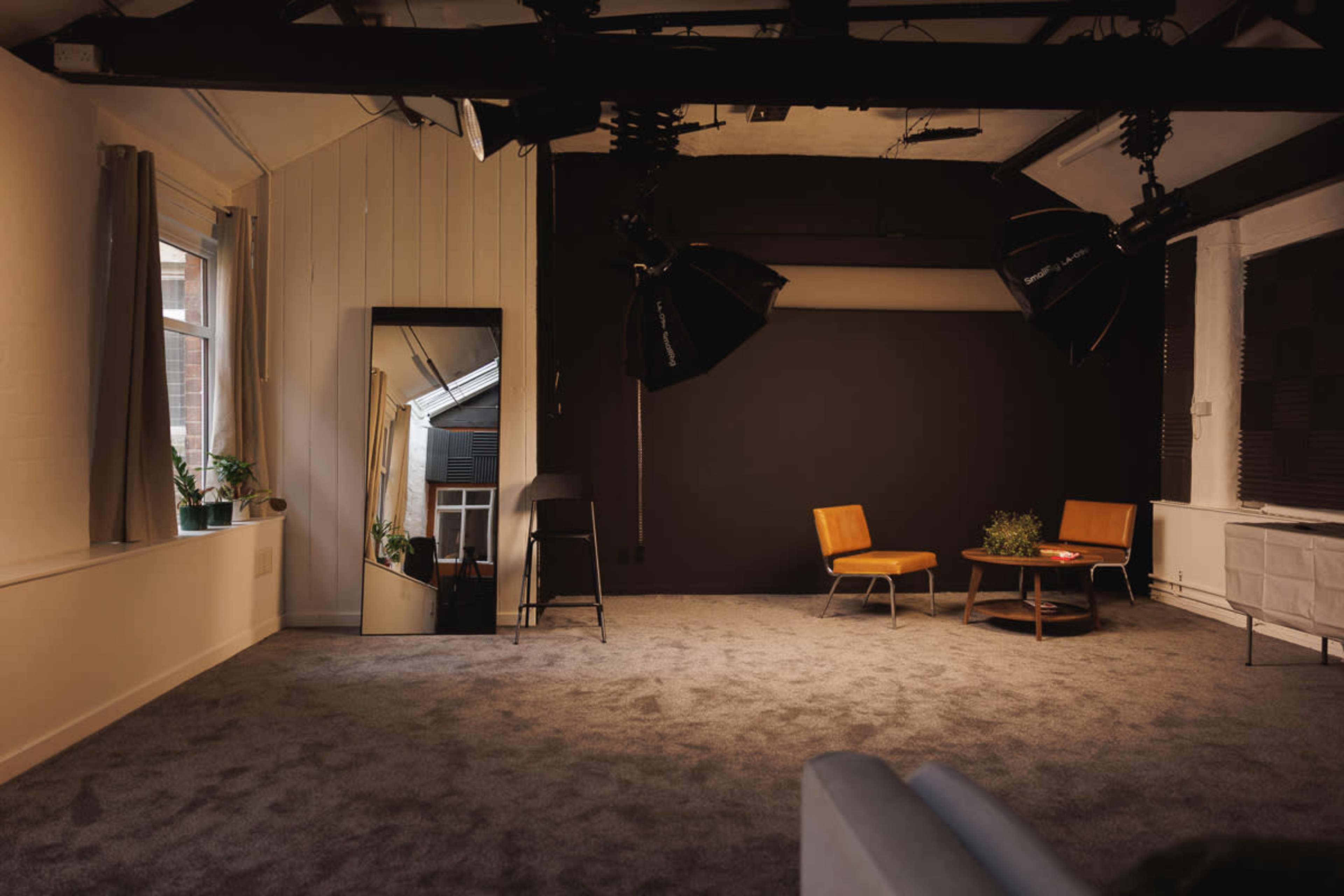 The image shows a spacious interior with gray carpeting, featuring two orange chairs, a low coffee table with a plant, a mirror, and large studio lights above.