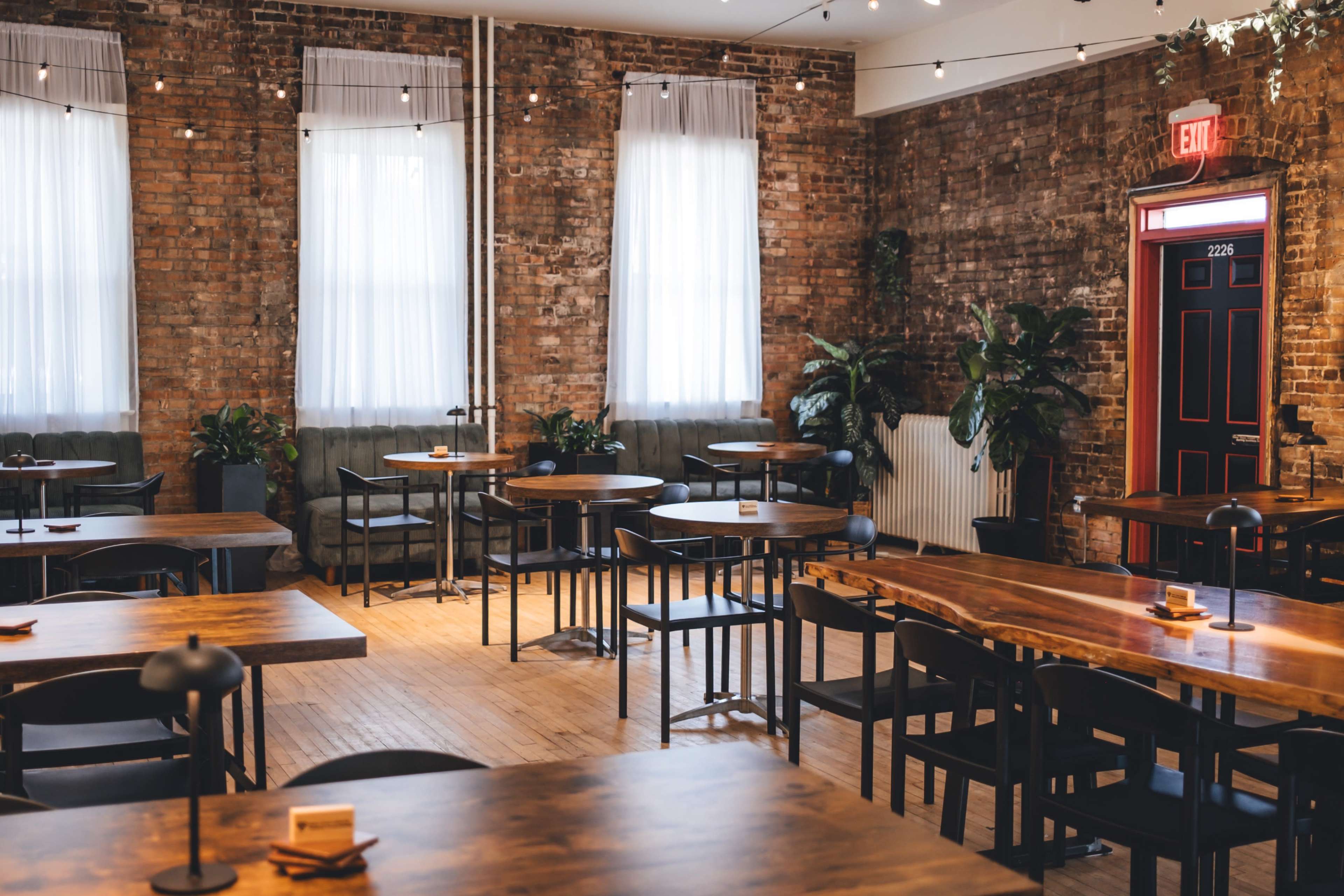 The image shows a warm-toned restaurant interior with wooden tables, chairs, and large windows, featuring brick walls and indoor plants.