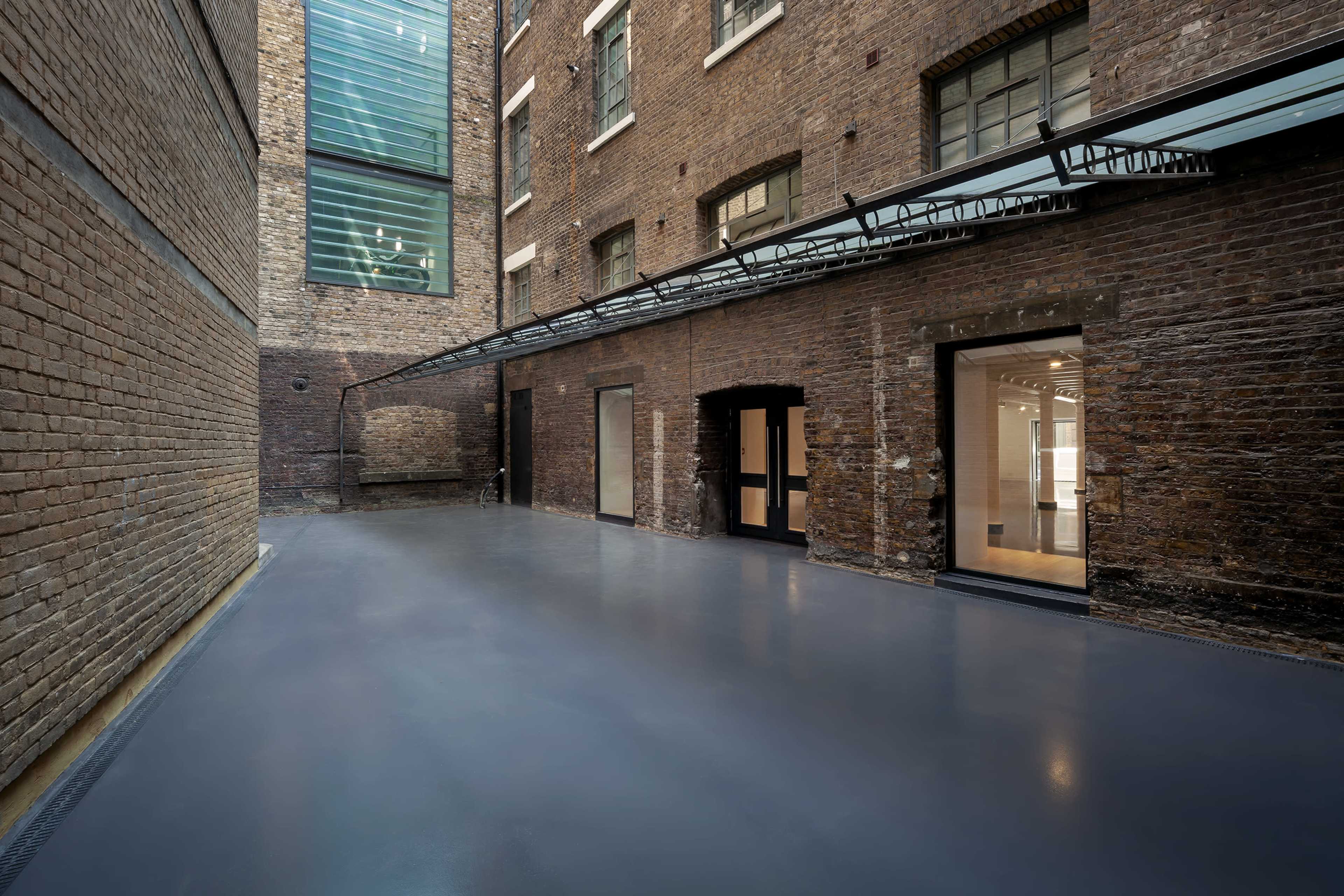 A narrow courtyard surrounded by brick walls and large windows, with a smooth, gray floor and entrances to adjacent rooms.