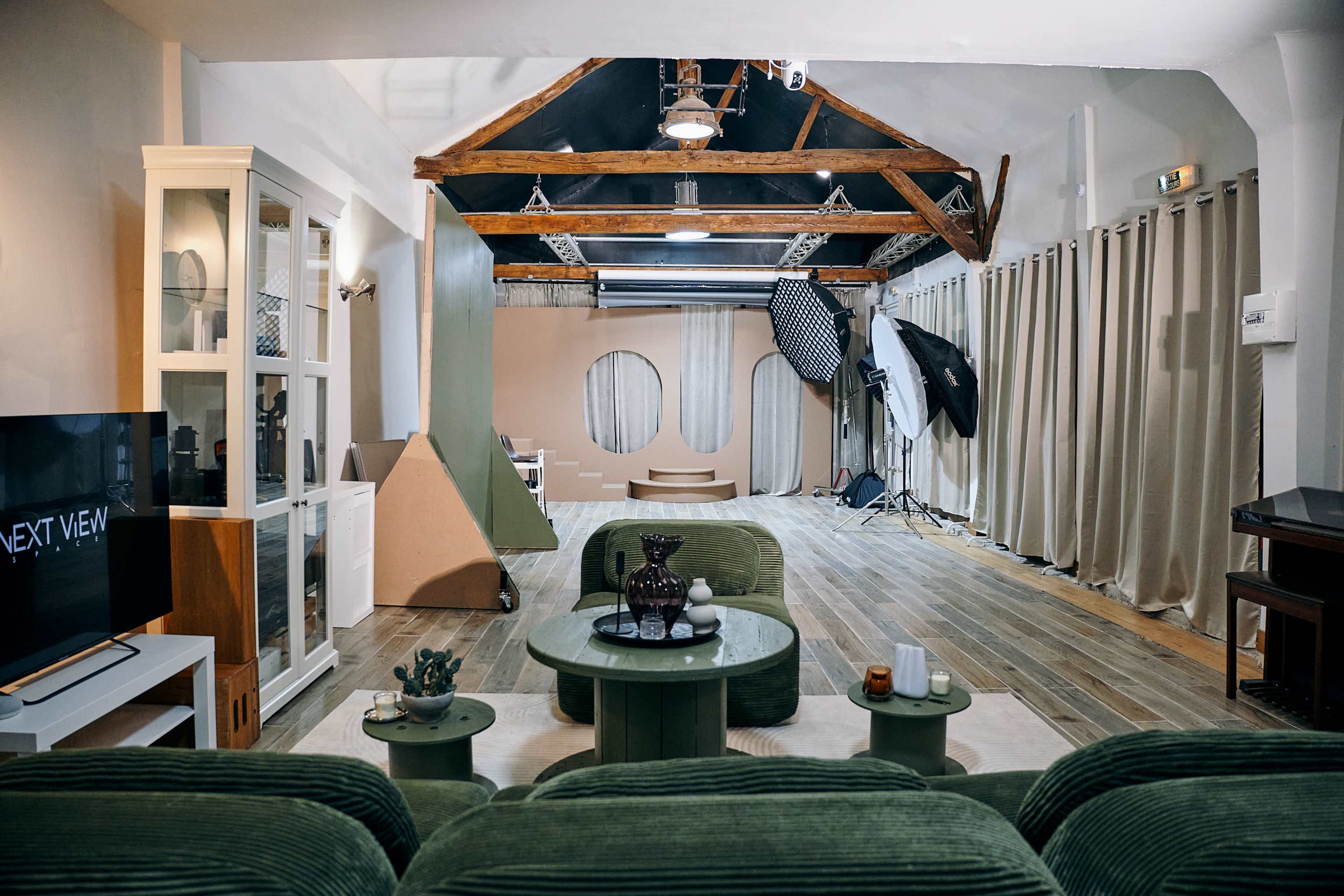 The image shows a spacious interior with wooden beams, a light-colored floor, green upholstered furniture, and a minimalist design featuring decorative elements and photography equipment in the background.